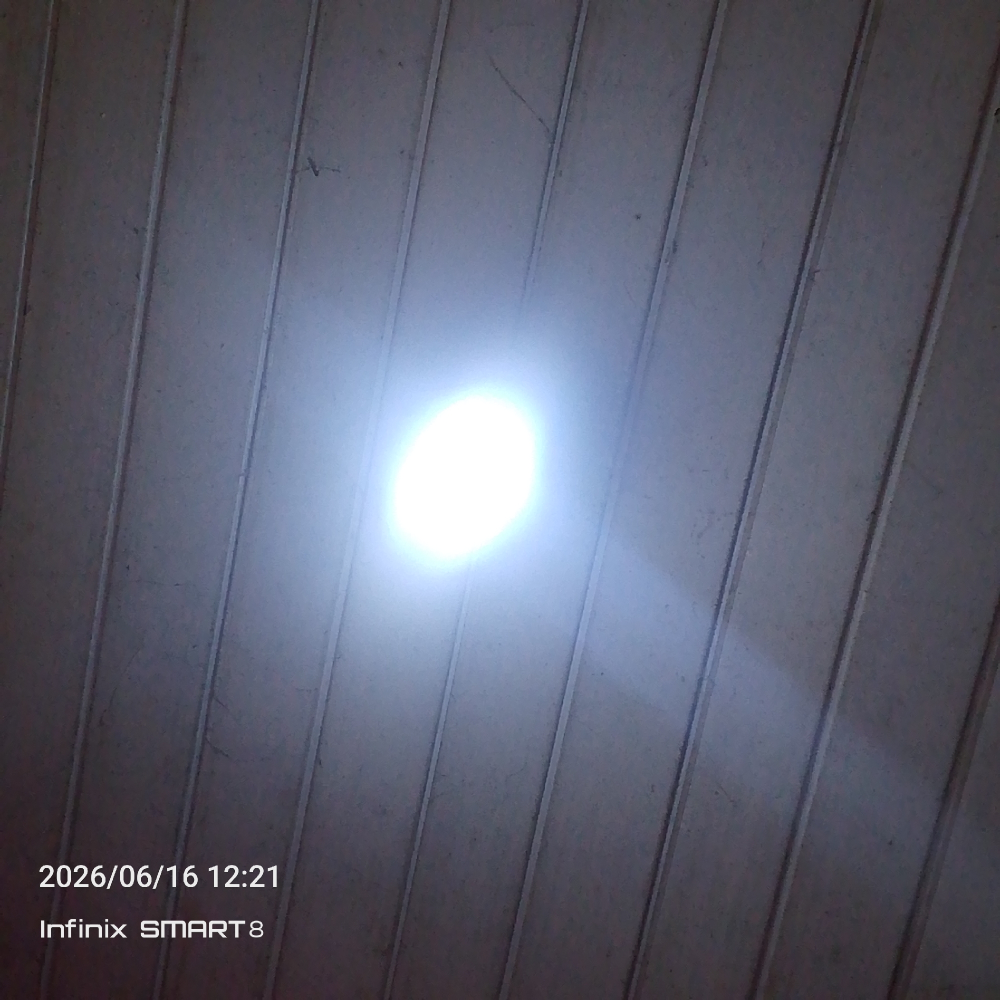 The image is a ceiling with vertical lines, possibly a tin ceiling or paneling. A bright light source is centrally located, creating an oval-shaped illumination with a halo effect. The light is so intense that it washes out detail in its immediate vicinity. The overall color palette is muted grays and whites, with subtle shadows cast by the ridges in the ceiling. In the bottom left corner, there is text overlay that reads "2026/06/16 12:21 Infinix SMART 8". The scene appears to be indoors, with no indication of an outdoor environment or specific city.