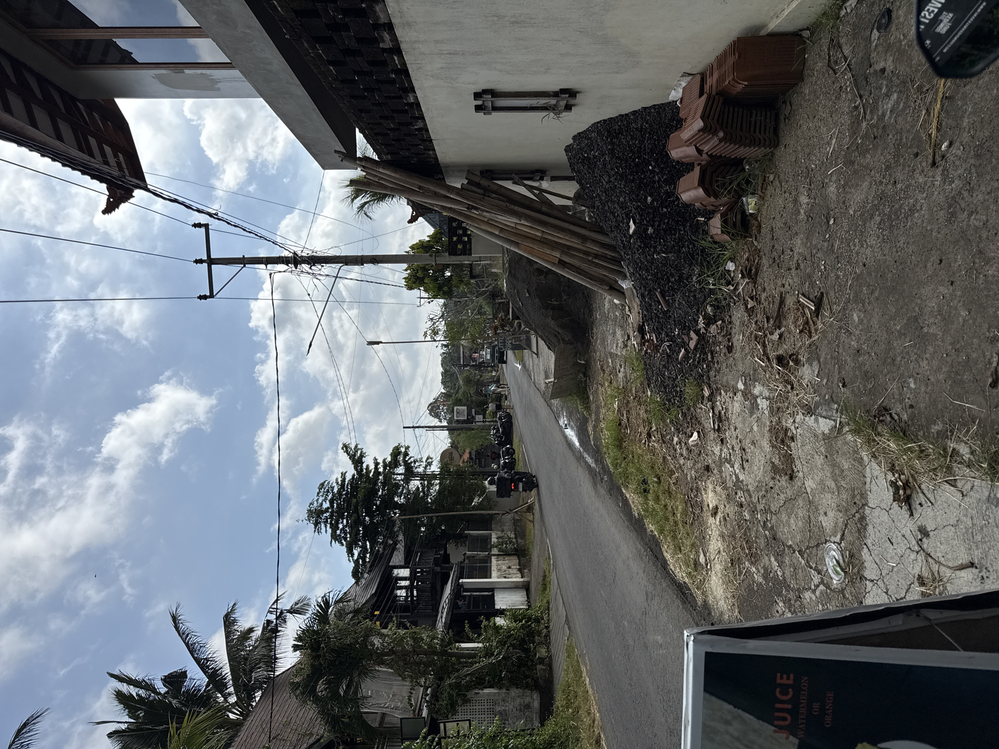 The image depicts a narrow asphalt road in an urban environment, likely in a tropical or subtropical location. The road curves gently into the distance, bordered by buildings and vegetation.
On the right side of the road, there's a building with a white facade and exposed wooden beams. Construction materials, including bamboo poles, a pile of black gravel-like material, and stacks of terracotta roof tiles, are stacked against the building. The road edge is cracked and overgrown with grass and weeds. A discarded aluminum can is visible on the ground, and a corner of a sign with the word "JUICE" in a large font is visible in the foreground.
On the left side of the road, buildings with traditional Balinese architectural elements, including thatched roofs and wooden details, are visible. The scene includes palm trees and other lush vegetation. Overhead, utility poles and power lines crisscross the sky, which is partly obscured by clouds. A line of parked motorcycles are visible along the roadside in the distance. One motorcyclist is driving on the road.