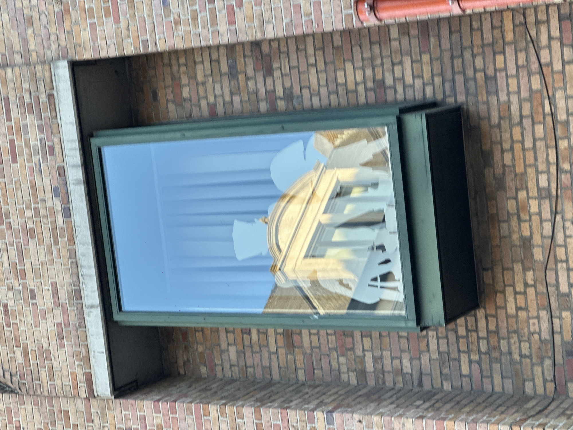 The image shows a close-up, exterior view of a window in Bruges, Belgium. The window is framed by a dark green border and is set into a brick wall made of light brown and reddish bricks. Inside the window, there appears to be a display or artwork created with white paper cutouts. These cutouts suggest figures or architectural elements, with one cutout resembling a top hat and another possibly a building facade with columns. The reflection in the glass shows a light blue sky and some architectural details, possibly of buildings opposite. Below the window, there is a dark green, box-like structure. A bright orange pipe runs vertically along the right side of the brick wall.