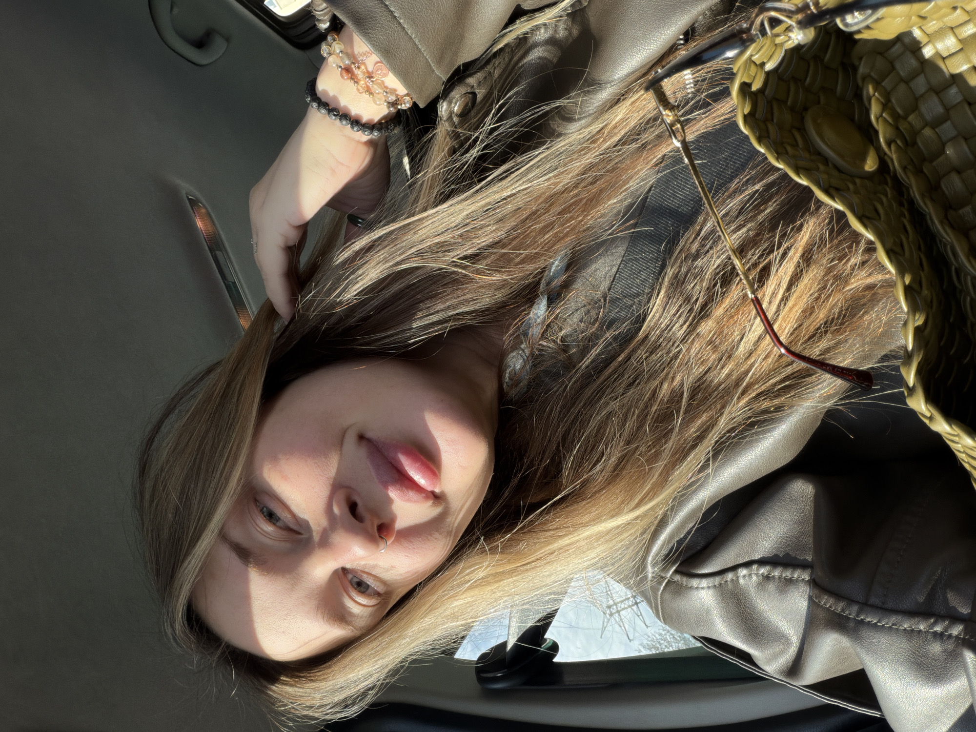 A young person with long, blonde to light brown hair is depicted in a close-up selfie taken inside a car, with the image rotated 90 degrees clockwise. They have light-colored eyes and a septum piercing, with their lips slightly parted in a relaxed expression. Strong natural sunlight illuminates the left side of their face, casting a distinct shadow across the right.
The person is wearing a brown or tan leather-like jacket, with its texture and stitching visible. On their left wrist, multiple bracelets are stacked, including one with black beads (possibly lava stone) and another with lighter, translucent beads.
To the right of the frame, a partially visible olive green or khaki-colored woven bag is present, from which a pair of glasses hangs. Faint text "CO SP" can be discerned on the arm of the glasses.
The setting is the interior of a car, evidenced by the gray headliner, a grab handle, and a ceiling light fixture in the background. Through a window visible on the bottom right, hints of the outside environment, including power lines or utility structures against a bright sky, suggest a daytime scene. The person's hair is spread out, catching the light.
Location context: Chaikovsky, Russia.