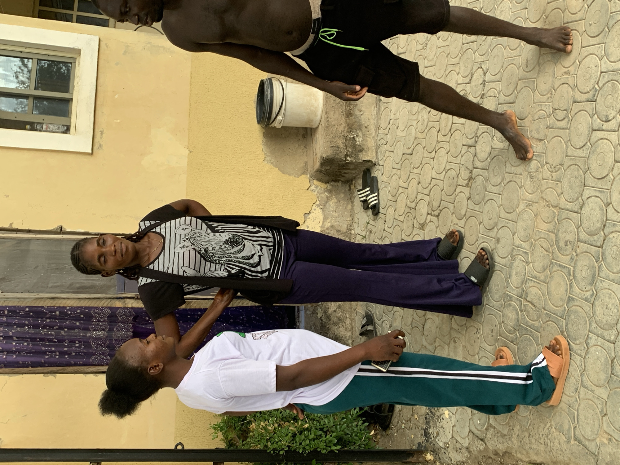 Three individuals are standing on a paved outdoor area in front of a building. The building has a yellow-painted textured wall with a white-trimmed window. A dark-colored door with a purple patterned curtain is visible to the left of the window.
On the left, a person wearing a white t-shirt and green pants with white stripes is facing towards the center, talking to another person. They have their hair in a bun. The person in the middle, a woman, is wearing a black and white striped t-shirt with a zebra graphic and dark purple bell-bottom pants. She is facing towards the viewer and has braids. Her left hand is on the hip of the person in the white t-shirt.
On the right, a shirtless person wearing black shorts with a bright green drawstring is standing, facing to the left and looking towards the other two individuals. Their legs are slightly spread apart.
In the background, a white bucket with a black lid is placed on a concrete block. Several pairs of sandals are visible on the paved ground. A green plant is growing to the left of the building. The overall scene appears to be a casual outdoor gathering or interaction in Abuja, Nigeria.
