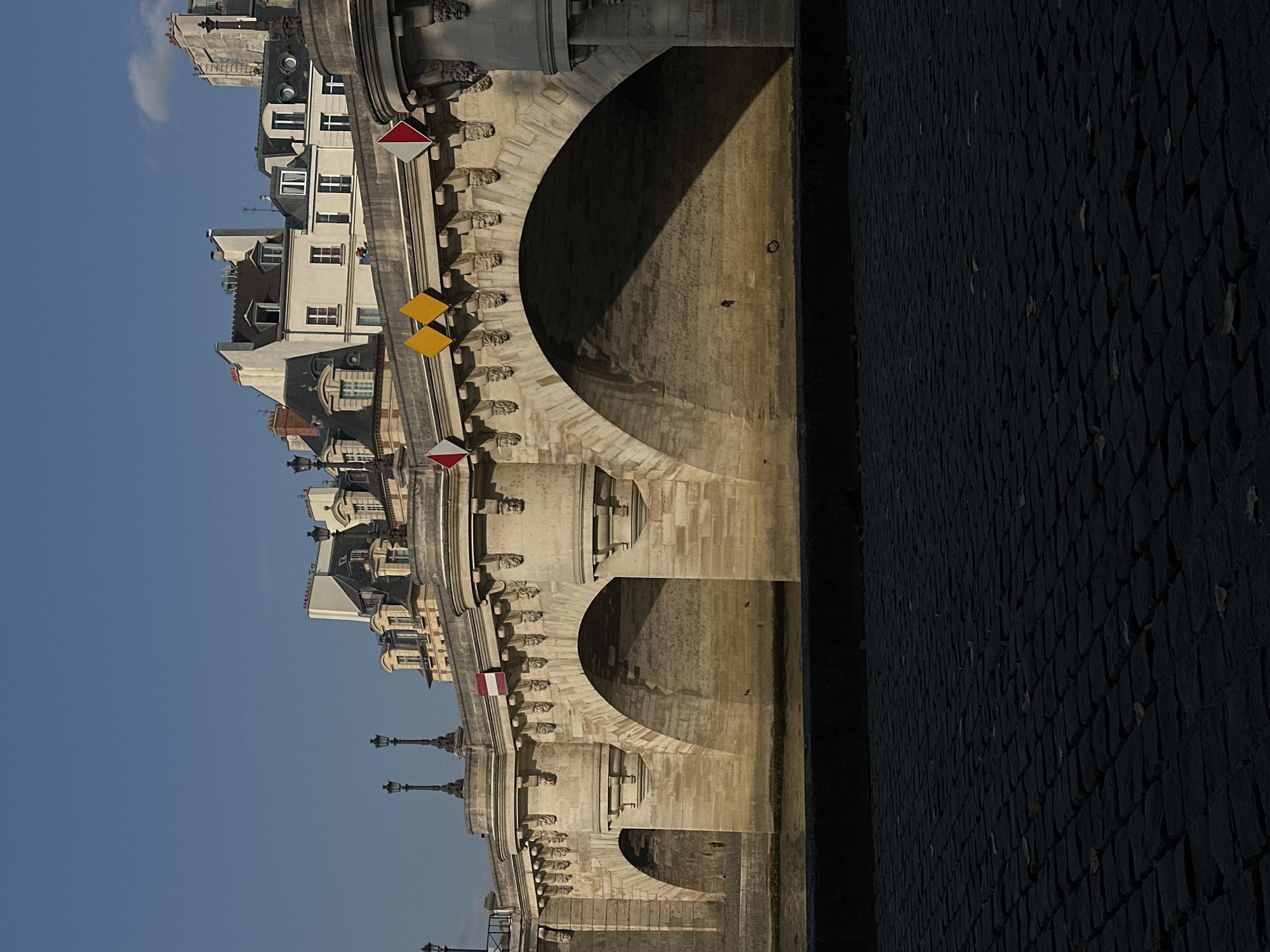 The image shows a stone bridge with multiple arches spanning a body of water. The bridge is adorned with ornate carvings and is topped with buildings with distinctive Parisian architecture, including mansard roofs and dormer windows. Decorative lampposts line the top of the bridge. Small diamond-shaped signs, some red and white, and some yellow, are attached to the bridge's facade. The foreground is paved with dark cobblestones, and some scattered leaves suggest an autumn or early winter setting. The sky is a clear, bright blue. The scene is captured in natural daylight, with strong shadows indicating a low sun angle. The overall impression is one of historical grandeur within an urban environment.