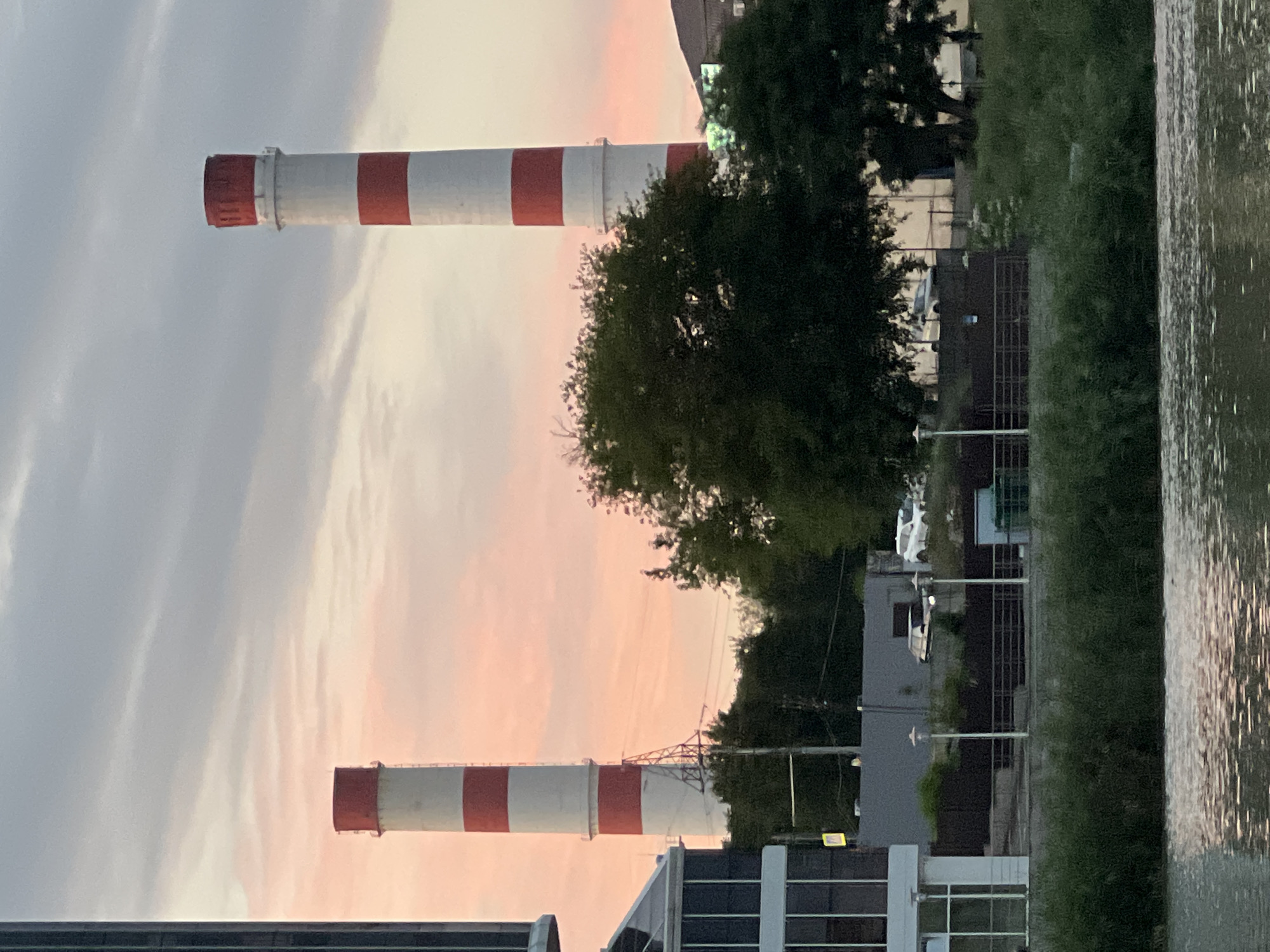This media file depicts an industrial scene at dusk, with two tall smokestacks, painted with red and white stripes, dominating the frame. The smokestacks are the primary objects, suggesting a factory or power plant setting. They are positioned vertically in the frame, with the lower one closer to the foreground and the upper one further back.
Behind the smokestacks, a dense cluster of dark green trees is visible, partially obscuring buildings. The architecture of the buildings appears modern, with some featuring large windows. The sky is a prominent feature, displaying soft, pastel hues of pink and grey, indicative of a sunset. This time of day contributes to a calm and serene atmosphere, contrasting with the industrial nature of the smokestacks.
To the right of the frame, a body of water is present, with its surface reflecting the ambient light, creating shimmering patterns. The presence of water suggests the industrial site might be located near a river or canal. The overall scene conveys a sense of industrial activity harmonizing with nature during a peaceful evening. There are no people or discernible text visible in the image. The weather appears to be clear, with the soft lighting suggesting fair conditions. The location context of Krasnodar, Russia, provides a specific geographical anchor for this industrial landscape.