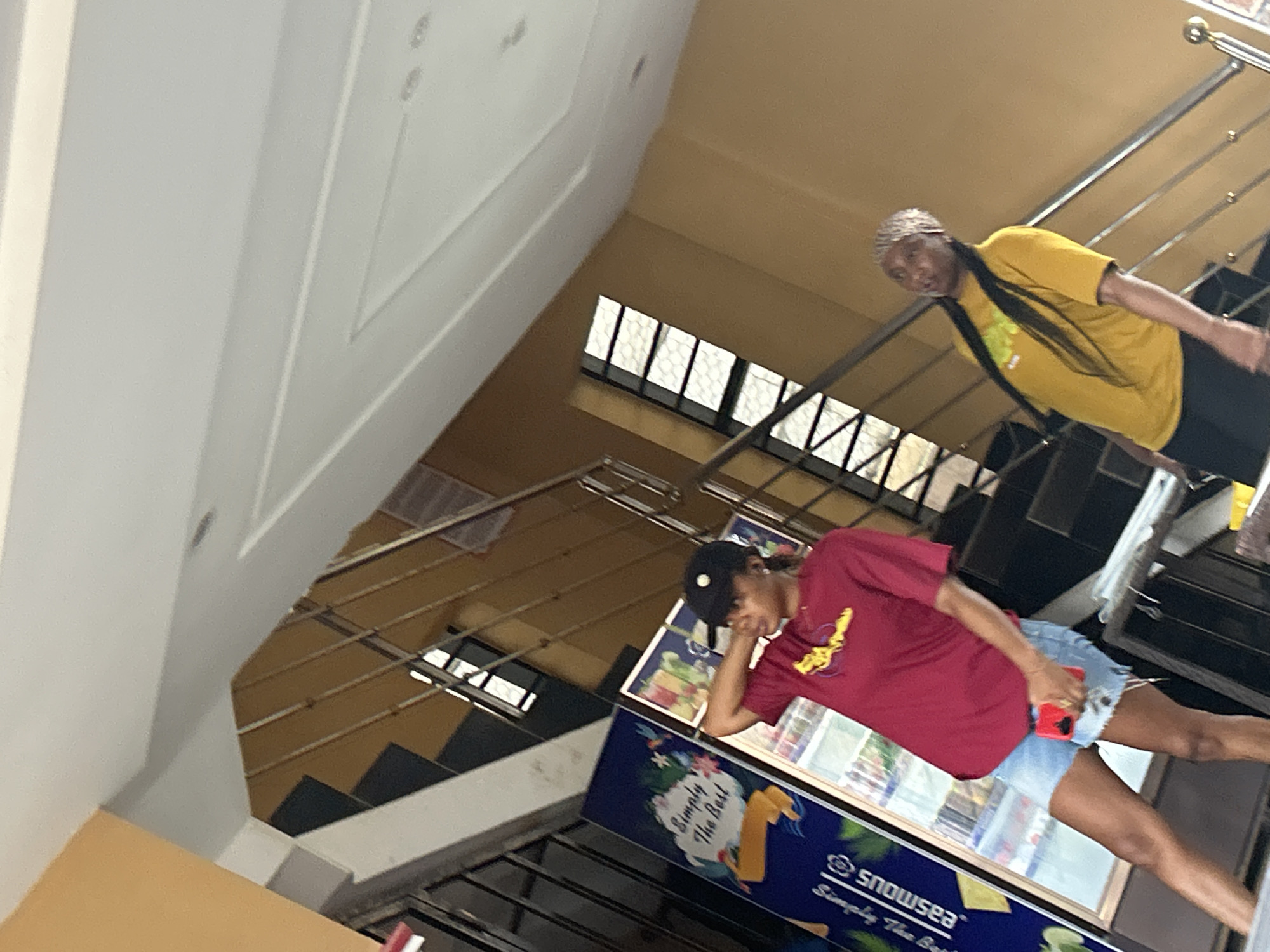 In Ihiagwa, Nigeria, a young woman in a red t-shirt and denim shorts, wearing a black baseball cap, stands near a refrigerator display. She holds a red smartphone in her right hand. To her right, another young woman in a yellow t-shirt and black pants, wearing a patterned headscarf and with long black hair, stands on a staircase. She looks towards the camera, her mouth slightly open. The background reveals a staircase with metal railings, leading upwards. The environment appears to be an indoor public space with tiled floors and beige walls, possibly a shopping center or a similar establishment.