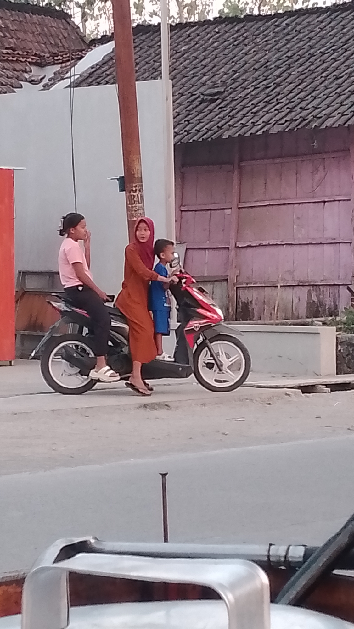 In Karanglangu, Indonesia, a motor scooter carries three individuals: a young boy in a blue shirt and shorts in front, a woman wearing a rust-colored dress and hijab behind him, and a girl in a pink shirt and black pants behind the woman. The motor scooter is parked on an asphalt road. In the background, a building with a gray tile roof and pink walls stands adjacent to the road. A utility pole is positioned in front of the building.