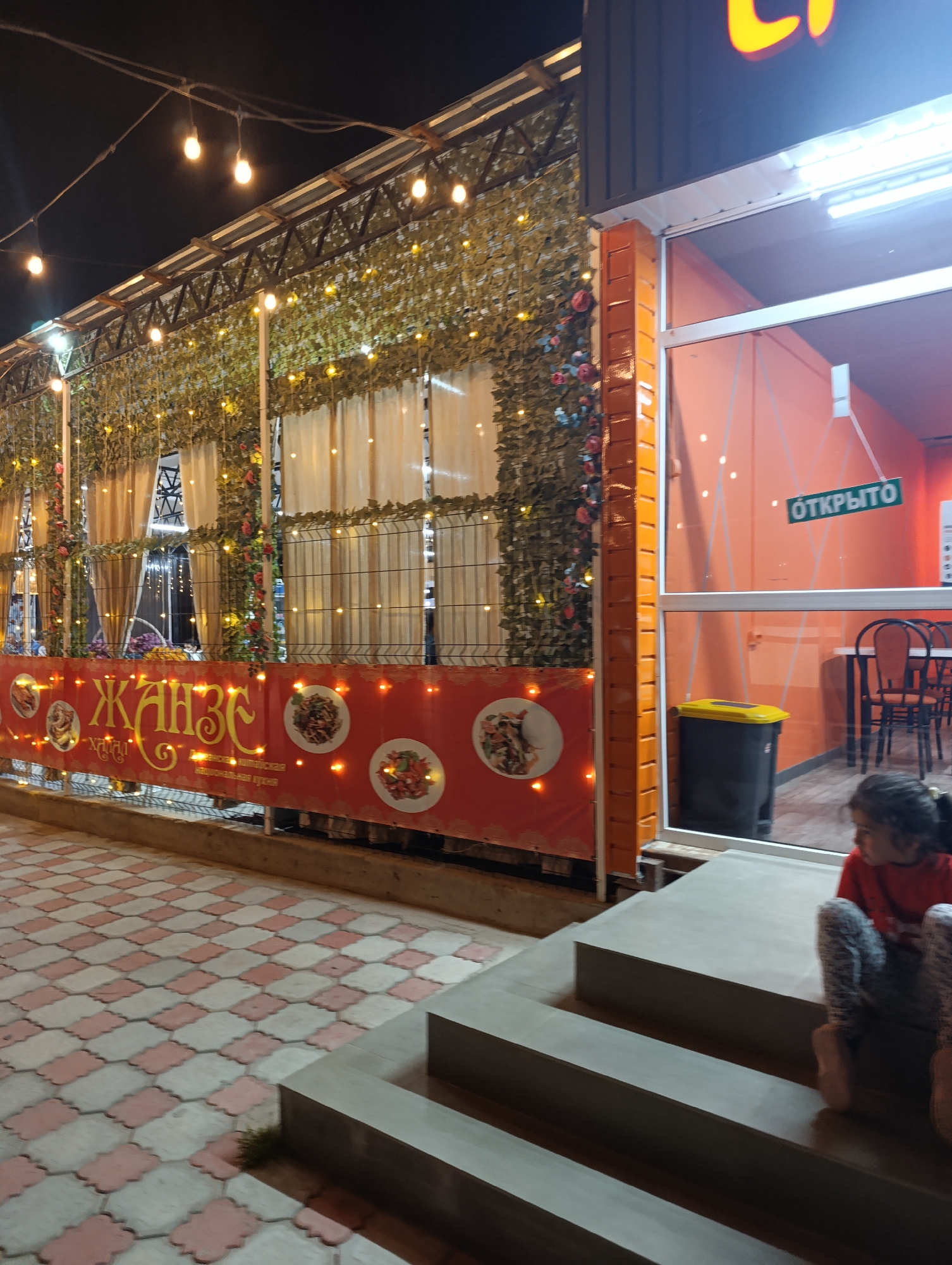 The image depicts the exterior of a restaurant or cafe at night in Bosteri, Kyrgyzstan. The facade features a red banner with text in Cyrillic script, displaying images of food dishes. Above the banner, decorative green foliage and fairy lights are visible, draped over a fence and roof structure. The building itself has an orange-colored exterior wall and a large glass window. Inside the window, tables and chairs are visible, along with a green sign that reads "ОТКРЫТО" (Open). A black trash can with a yellow lid is situated outside the window. In the foreground, a young girl wearing a red shirt and patterned leggings is seated on a series of concrete steps. The ground is paved with interlocking hexagonal tiles in red and off-white. The overall scene is illuminated by artificial lights, creating a festive and inviting atmosphere.