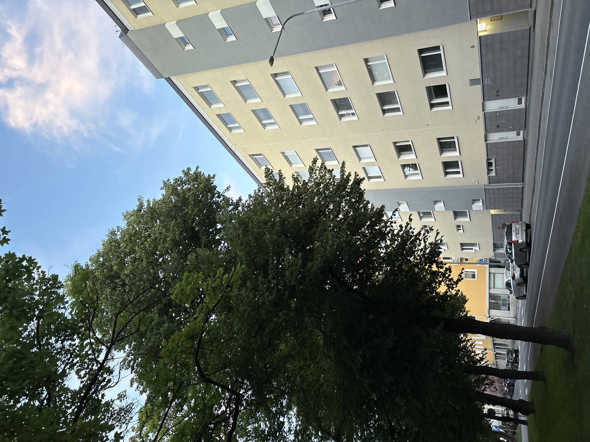 The image shows a residential apartment building in Pori, Finland, with a street and trees in the foreground. The building is tall with multiple stories, featuring a modern design with yellow and gray facades and numerous windows. A streetlamp stands to the right of the building. The street in front of the building has several cars parked and driving on it, including a silver sedan and a silver hatchback. Trees with green foliage line the left side of the street, casting shadows on the road and sidewalk. The sky above is blue with scattered clouds.