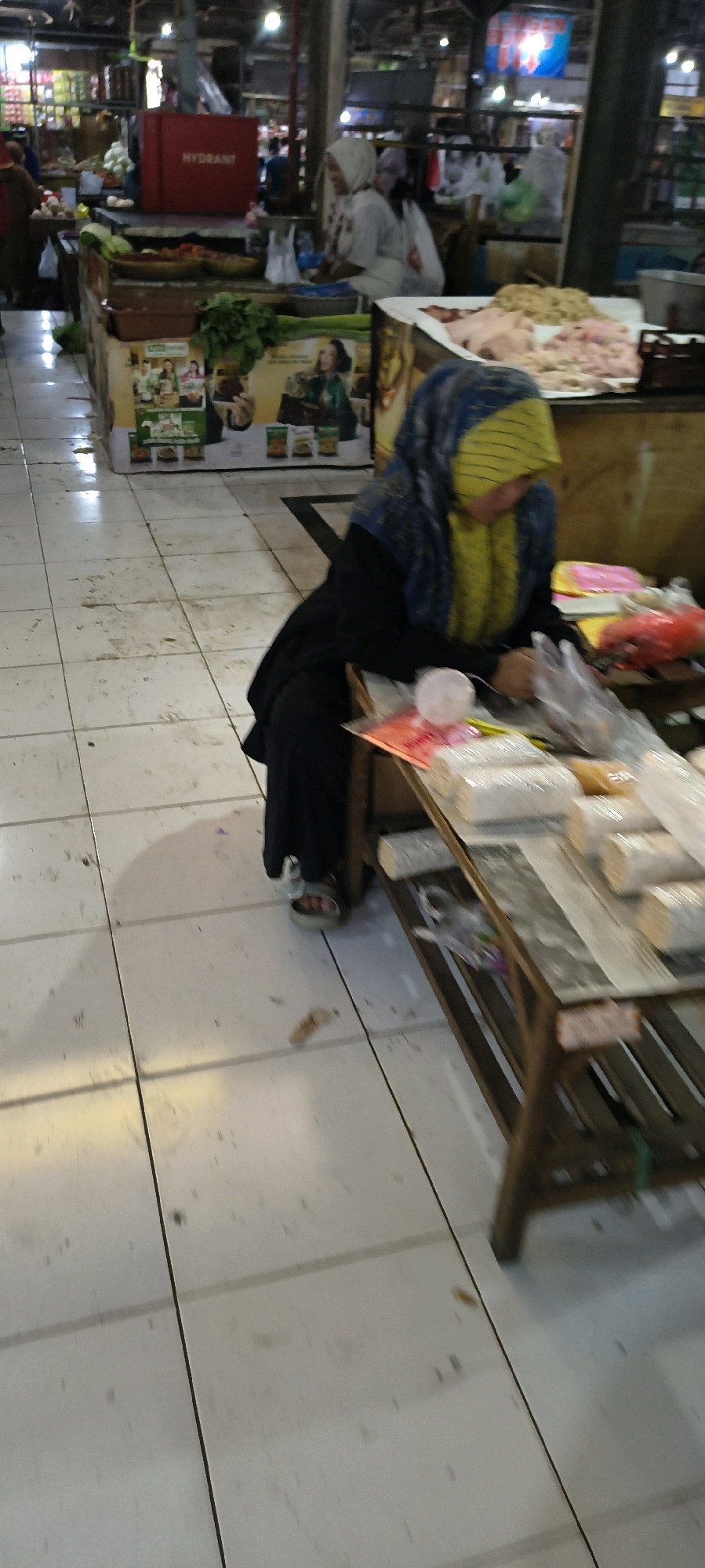 The image captures an indoor market scene in Weru Lor, Indonesia, characterized by a tiled floor and overhead lighting. In the foreground, a woman wearing a blue and yellow hijab and a black abaya is seated at a wooden stall. She is looking down and appears to be engaged in an activity involving plastic-wrapped goods on the stall's surface, which also displays loose food items and red plastic bags. To the left, a red fire hydrant box with white lettering that reads "HYDRANT" is prominently displayed on a counter filled with produce. In the background, other market stalls and individuals are visible, along with advertisements and various goods for sale. The overall ambiance suggests a bustling marketplace.