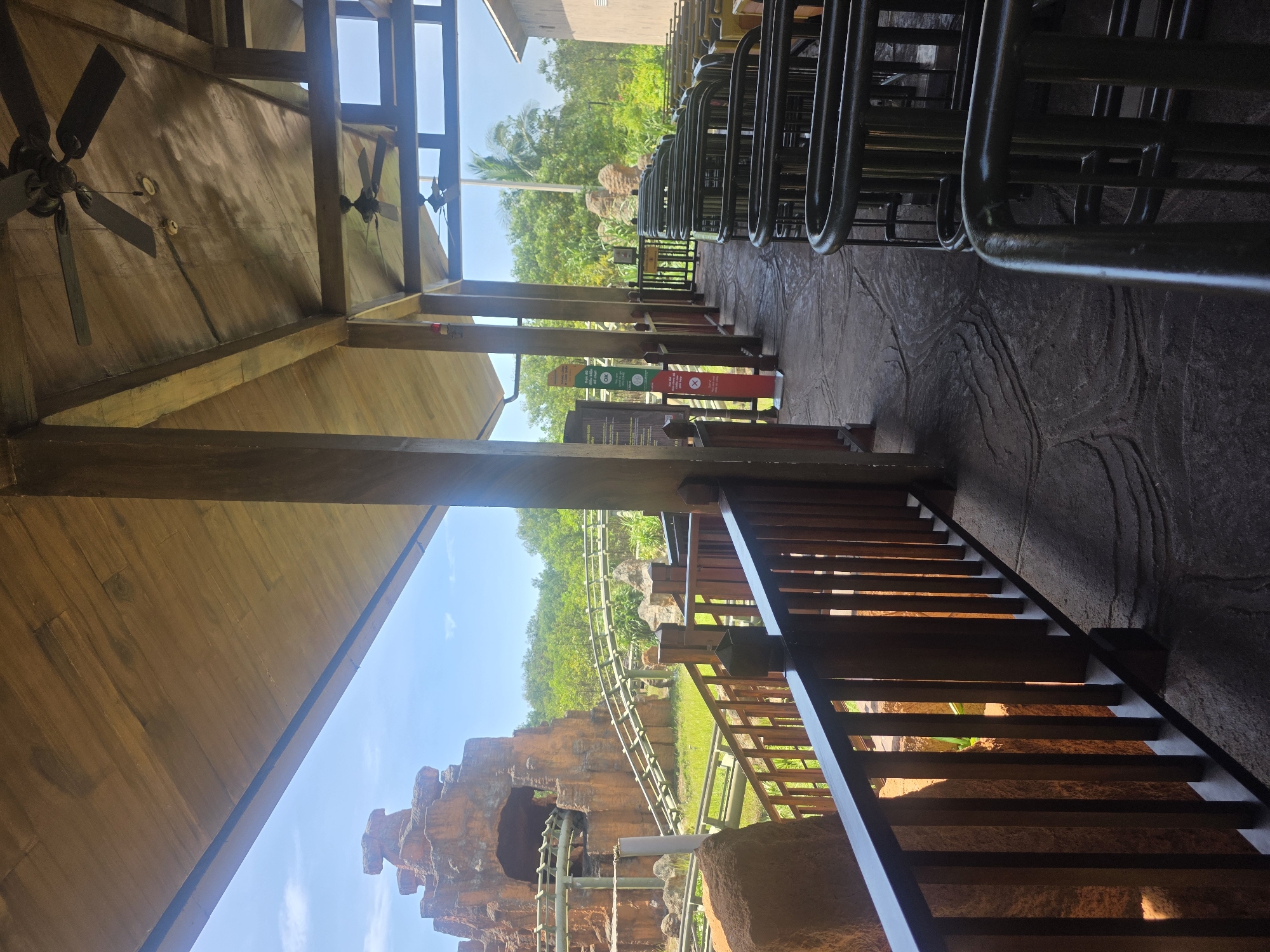 The image captures an indoor/outdoor waiting area for a roller coaster ride. The view is slightly tilted, oriented towards the roller coaster track which winds through a landscape of rock formations and greenery, under a partly cloudy sky. The waiting area is covered by a wooden roof, with ceiling fans visible. A metal railing separates the waiting area from the track, with stairs leading down to what appears to be a lower level. On the right, metal barriers delineate the queue lines. Signs containing green and red text and symbols are present along the queue. The scene is likely located in a theme park. Although I can see what appears to be the roller coaster track and waiting area, I cannot identify a specific location within Đà Nẵng, Vietnam.