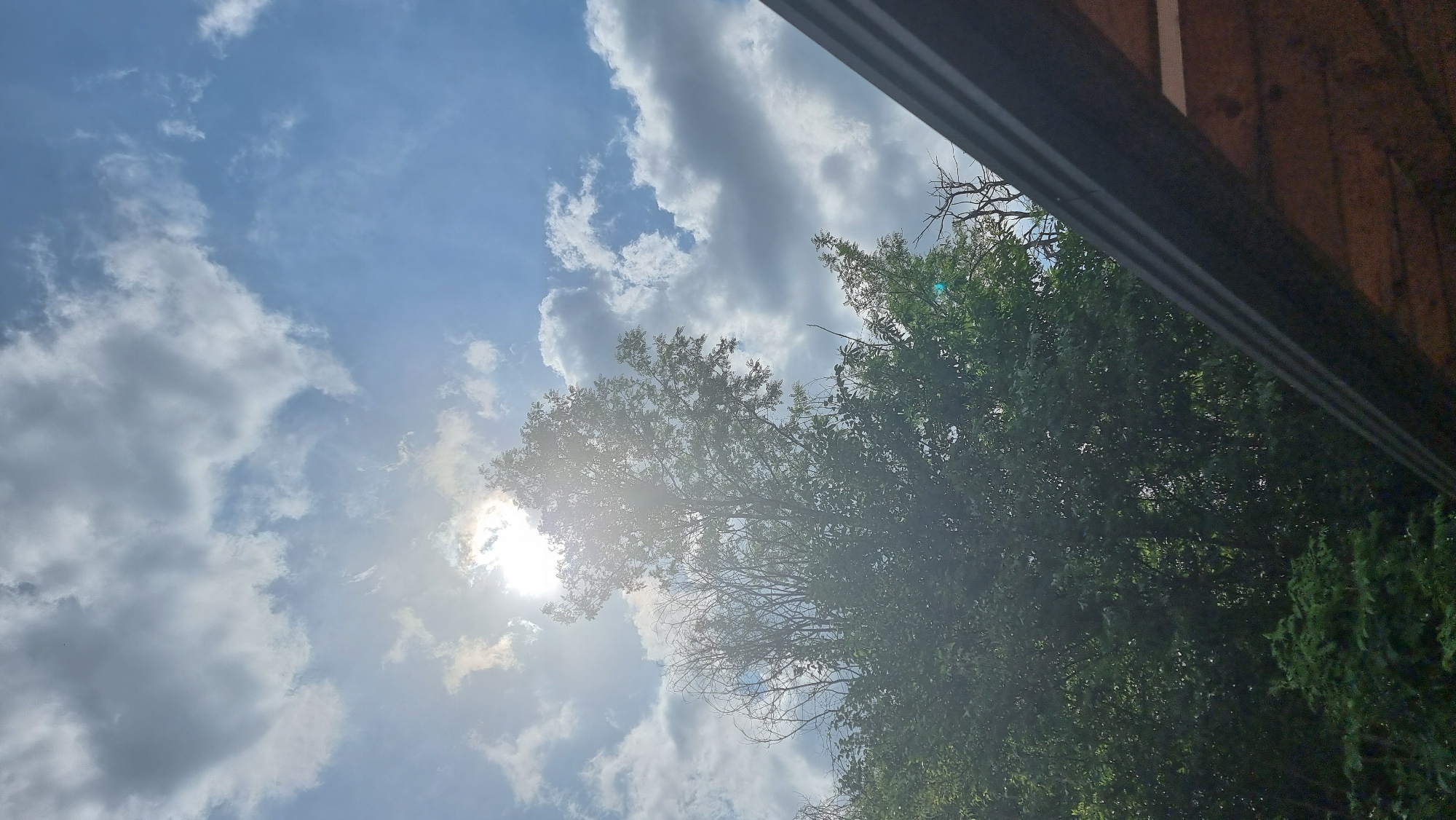 The image presents an upward view towards a partly cloudy sky, with sunlight filtering through tree canopies and a portion of a building structure. The sky, a clear blue in many areas, is adorned with soft white and grey cumulus clouds, indicating fair weather. The sun, exceptionally bright, is visible in the upper-left quadrant, partially obscured by clouds and tree leaves, creating a strong lens flare and radiating light.
Dominating the lower half and extending into the left side of the frame are dense, vibrant green foliage of several trees. Some areas also show bare, intricate branches silhouetted against the bright sky and sun, suggesting a mix of deciduous and evergreen trees, or perhaps parts of trees that are less leafy. The strong sunlight illuminates the leaves, giving them a slight glow.
In the upper-right portion of the image, a dark, horizontal architectural element, possibly a roof edge, gutter, or beam, cuts across the frame. Above it, lighter brown, wooden surfaces are visible, suggesting part of a building's eaves or exterior wall.
The scene captures a bright, sunny day, likely during midday or early afternoon, given the intensity and position of the sun. There is no specific activity or event taking place, and no people are visible. No text is discernable within the image. While the location context provided is Dunnae-myeon, South Korea, there are no specific visual cues within the photograph itself that indicate this geographical location.