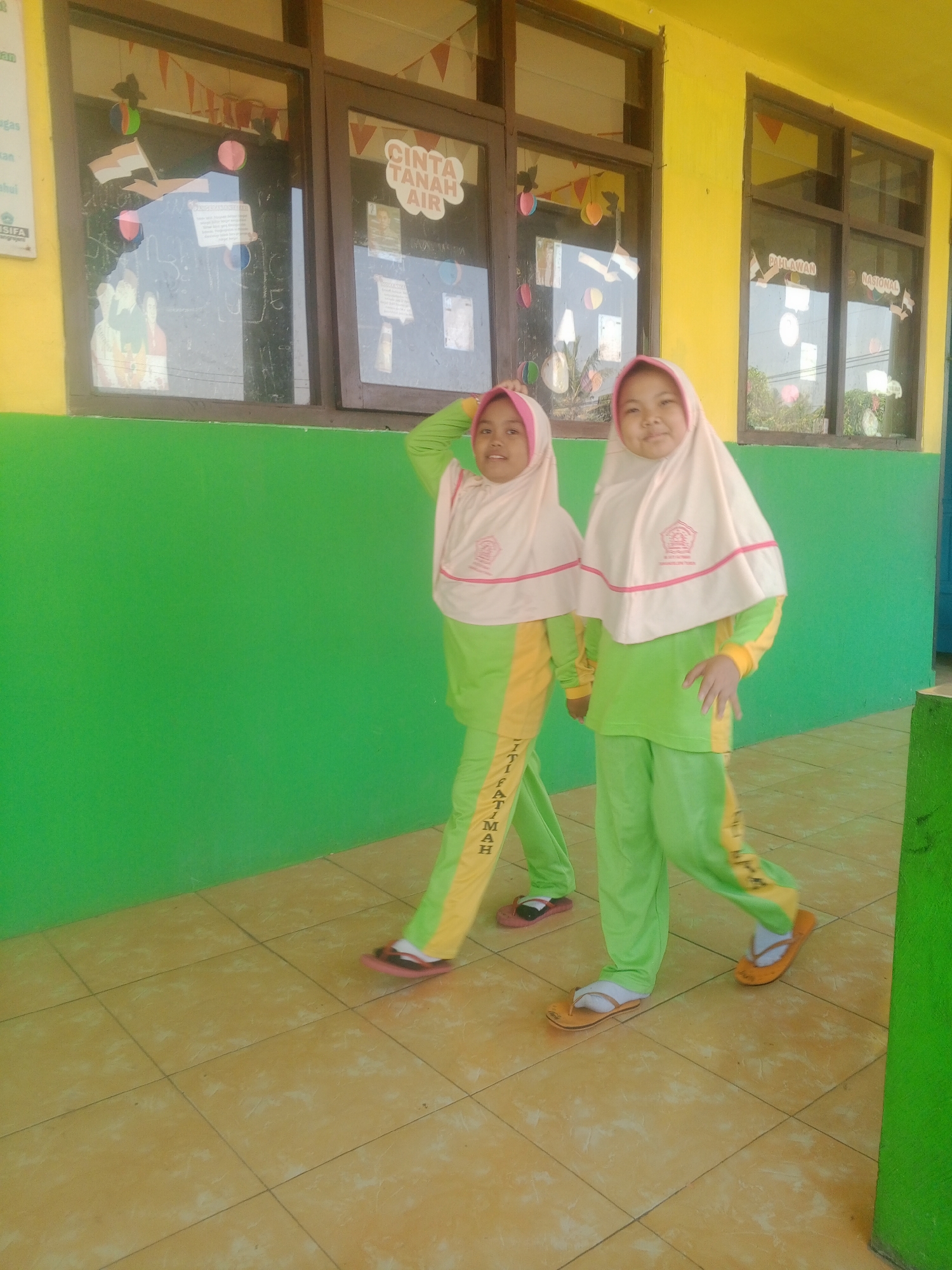 Two young girls, dressed in light pink hijabs and lime green tracksuits with yellow accents, stand on a tiled floor outside a building. The girl on the right smiles at the camera, while the girl on the left looks slightly away. The tracksuits have writing on the legs, partially visible as "JITI FATIMAH". The background features a bright yellow wall with brown-framed windows, through which a glimpse of trees and sky is visible. Red and white triangular flags are strung across the top of the windows. One window displays a sign with text that appears to be in Indonesian, including the phrase "CINTA TANAH AIR" (Love the Homeland). The floor is made of square, light brown tiles. The scene appears to be outdoors, possibly a schoolyard, in Kemulan, Indonesia.