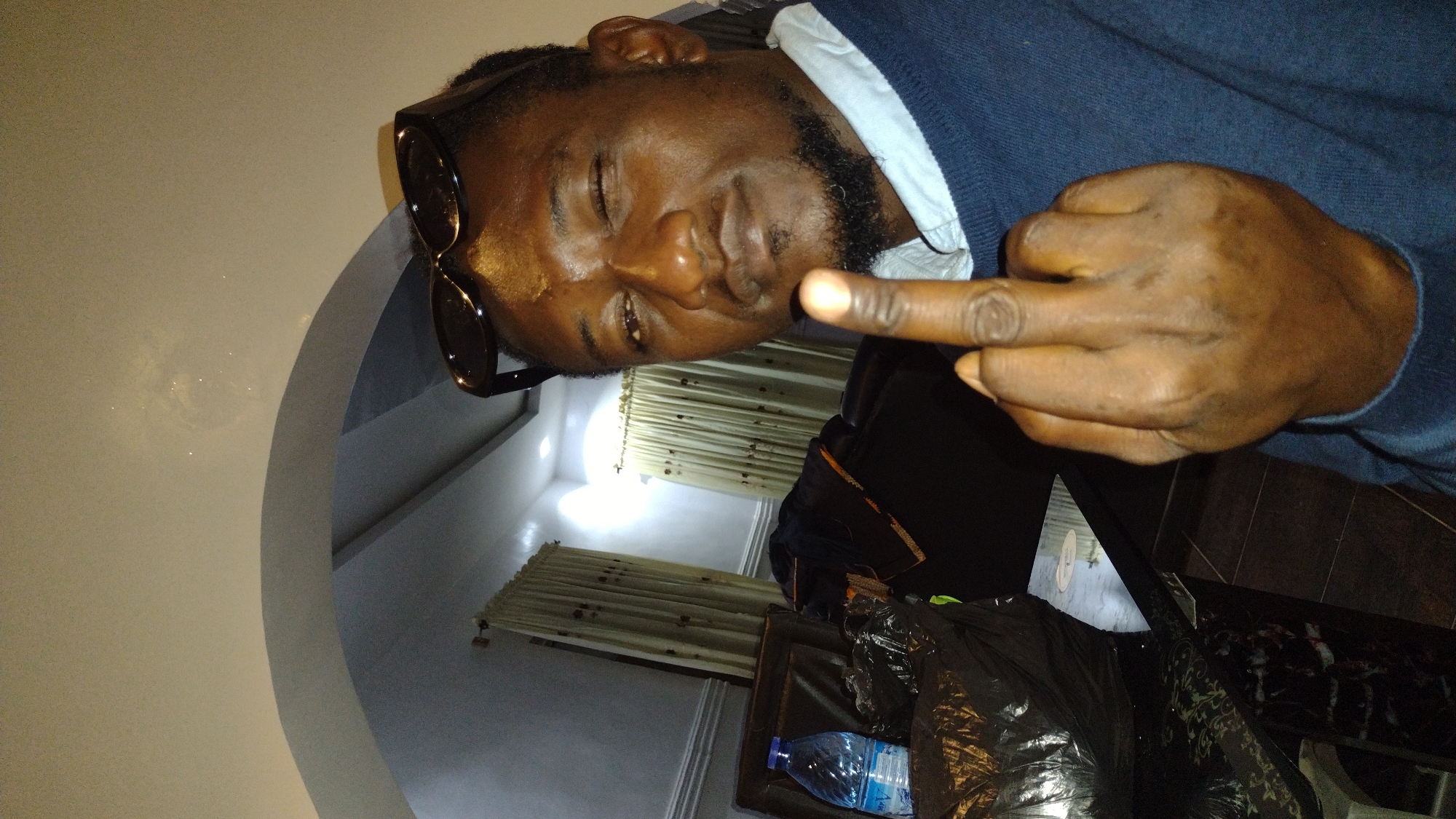 A close-up, low-angle selfie shows a Black man, appearing to be in his late 30s to early 40s, winking with his left eye. He wears dark sunglasses perched on his head, a light blue collared shirt under a navy blue sweater, and has a neatly trimmed beard and goatee. His right hand is raised towards the camera, with his middle finger extended in a gesture of defiance or obscenity. The background appears to be an indoor setting, possibly a living room in Jos, Nigeria. Curtains are visible, as well as a table with a bottle of water and some dark, crumpled items. The lighting is dim, creating shadows.