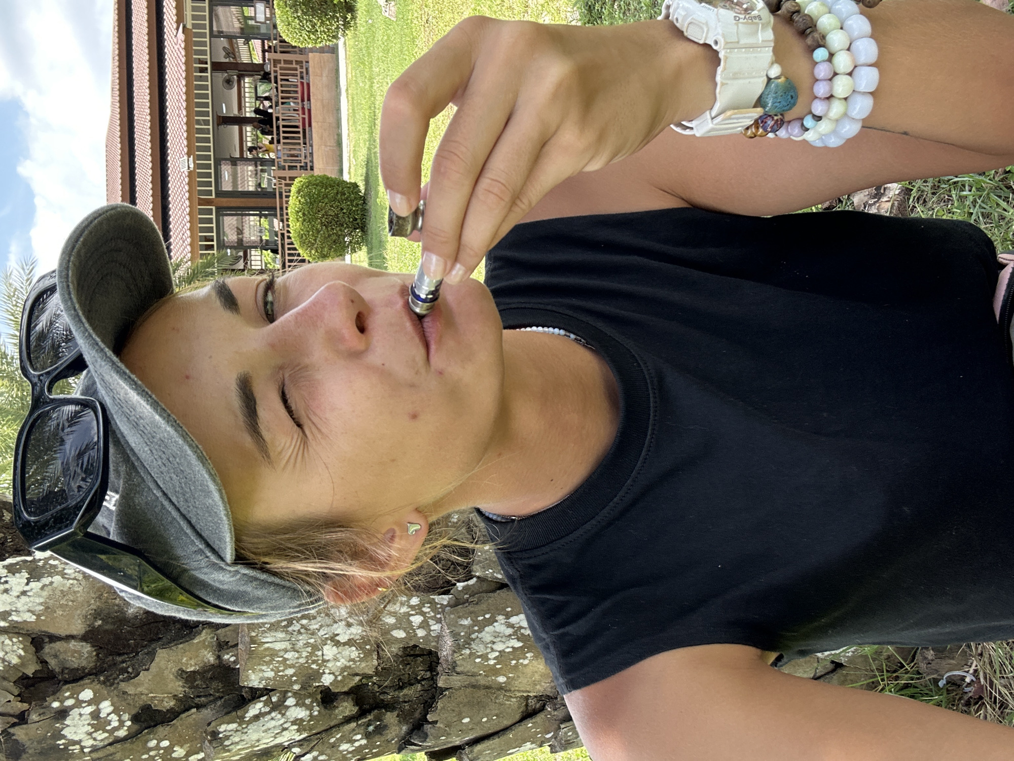 A woman is shown in a close-up outdoor shot, holding a smoking device to her mouth and winking. She is wearing a grey baseball cap with sunglasses perched on top, a black sleeveless shirt, and several bracelets on her right wrist. The background features a lush green lawn with a building and palm trees visible. The setting is the city of Ao Nang, Thailand, known for its beaches and tropical landscape. The lighting suggests it is daytime.
