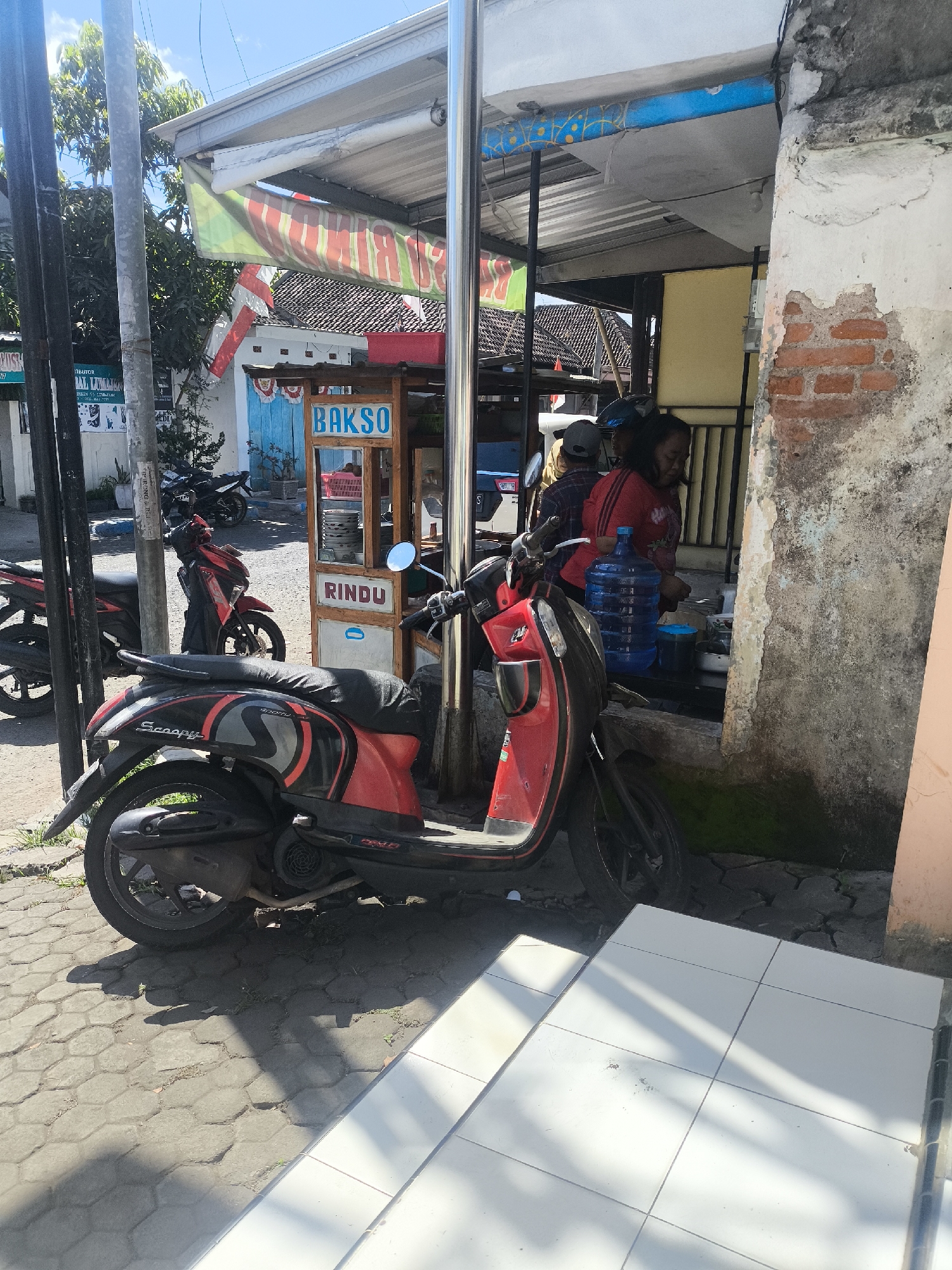 The image shows an outdoor scene in Lumajang, Indonesia, likely on a street or sidewalk. In the foreground, a red and black scooter, identified as a "Scoopy" by its branding, is parked. Behind the scooter, a food stall is visible with a sign that reads "BAKSO" (Indonesian for meatballs). A person wearing a red shirt is standing at the stall, behind a large blue water jug. Another person, wearing a grey cap, is partially visible. Various items, including bowls and what appears to be a cutting board, are on the counter of the stall. The stall has a wooden frame and a roof covered with a striped tarp. To the left of the stall, another red motorcycle is parked. The ground is paved with hexagonal stones, and there are some shadows cast by sunlight. In the background, there are trees and buildings, suggesting an urban environment. A street pole with multiple wires is also present on the left side of the image. The overall impression is of a street-side food vendor operating in a sunny, possibly warm, climate.