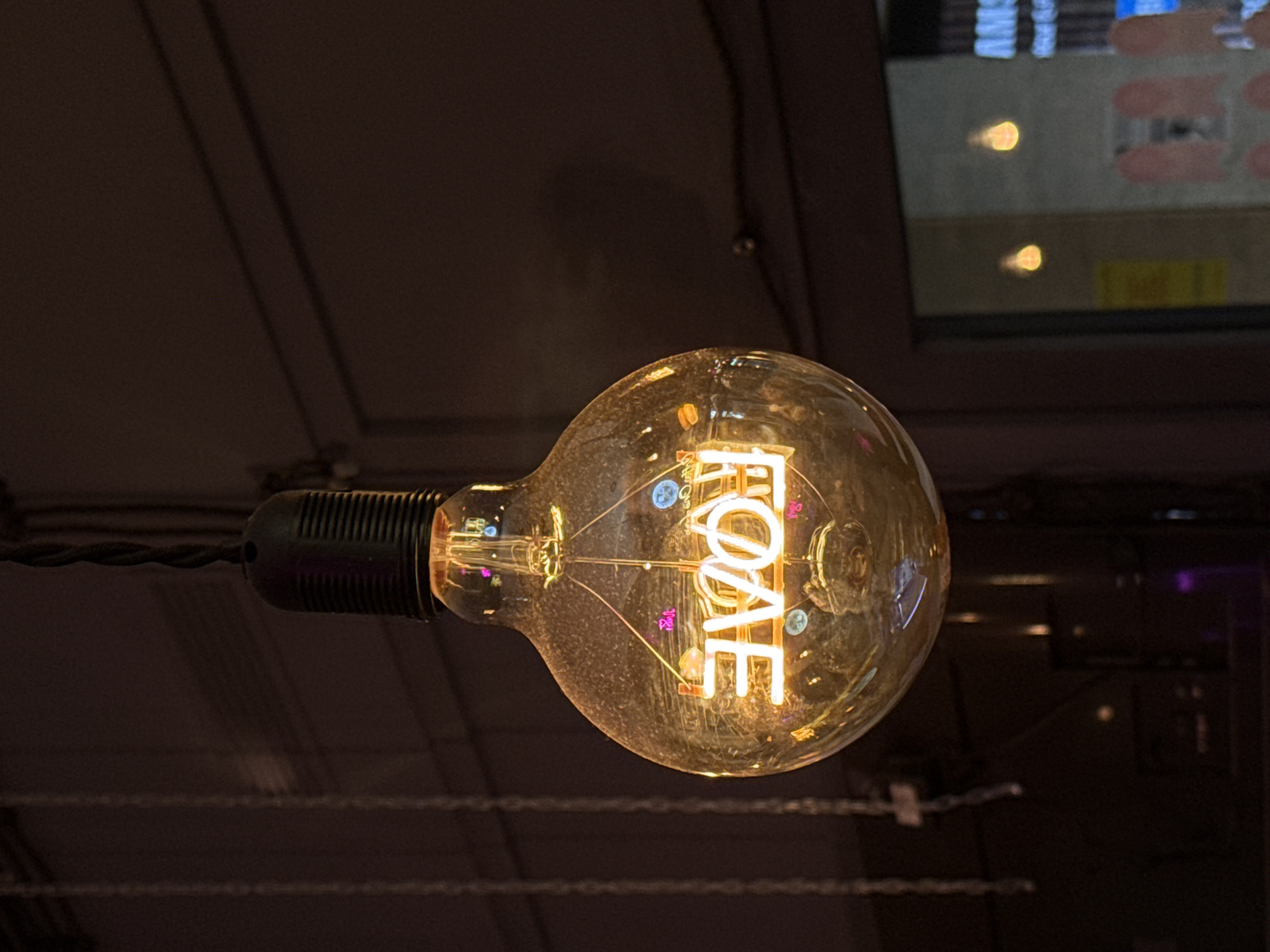 An illuminated Edison-style light bulb hangs from a black cord against a dark background. The bulb is made of clear glass and contains a neon sign that spells out "LOVE" in white light. The word is reflected in the glass of the bulb, appearing reversed. The background consists of a dark wall with some subtle lines and a hint of a window on the right side, showing blurred lights and text from an exterior environment. This is a detail shot of a decorative light fixture, with no specific identifiable landmarks suggesting a location in Moscow, Russia.