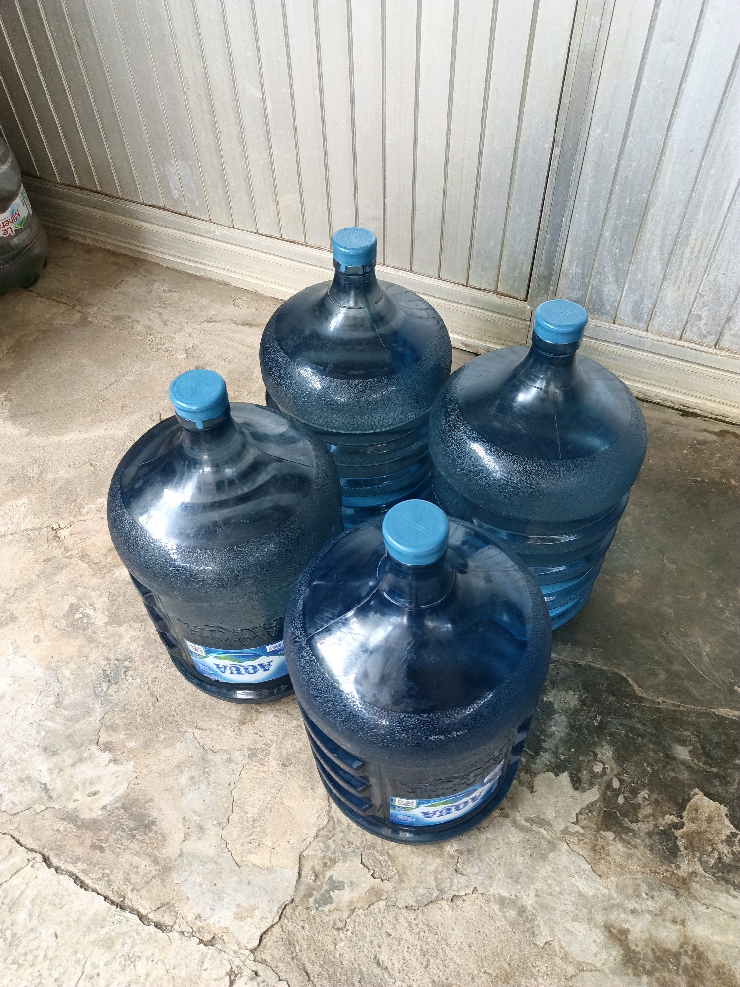Four blue, gallon-sized water jugs are placed on a cracked concrete surface in front of a corrugated metal wall. One jug is partially visible on the far left, and three jugs are clustered together in the center and right of the image. The jugs are all full, and the one in the foreground on the left has a label that reads "AQUA" in blue letters. The environment suggests an outdoor storage area, possibly behind a building or in a less-maintained part of a property. The lighting indicates it is daytime. The overall scene is static and does not depict any activity or interaction.
