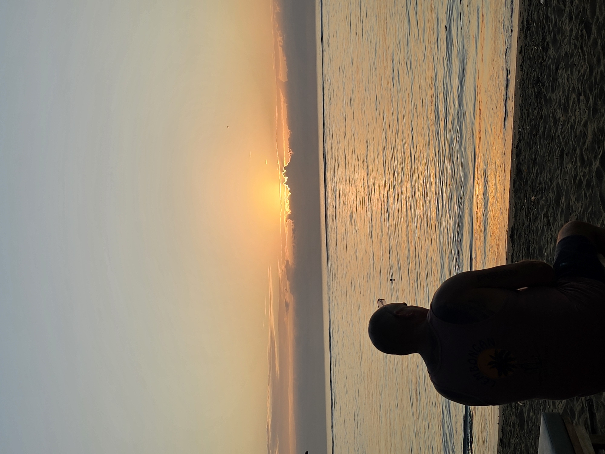 A man wearing a dark tank top sits facing away from the camera on a sandy beach in Denpasar, Indonesia. He is silhouetted against a tranquil ocean and a soft sunrise. The sun, partially obscured by clouds, casts a warm, orange glow across the water and sky. The man has tattoos on his arms and sunglasses resting on his head. In the distance, the calm sea meets the horizon under a gradient of pale blue and soft orange sky.