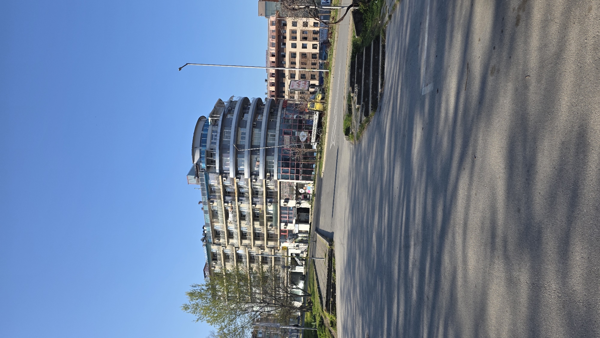 The image captures an urban scene in Blagoevgrad, Bulgaria, showcasing a distinctive curved apartment building with multiple balconies and glass panels. The building appears to be several stories high. The blue sky dominates the background, while the foreground features an asphalt road, marked with a white arrow to indicate the direction of traffic. On the right side of the road, concrete structures stand parallel to the road. Tree shadows fall onto the road.