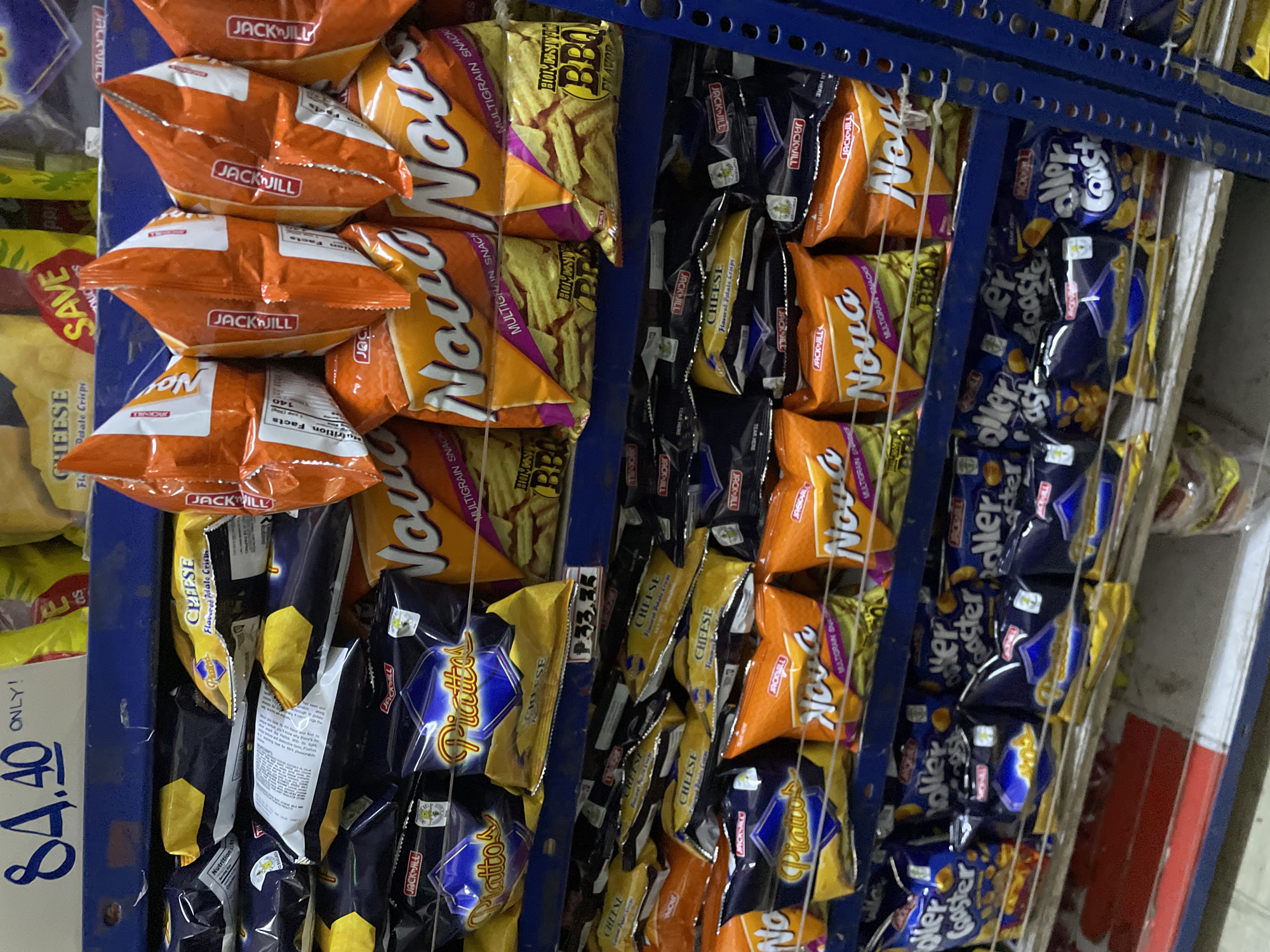 The image displays a retail snack food aisle within an establishment in Davao City, Philippines. A blue metal shelving unit occupies the majority of the frame, laden with various snack bags.
The top visible shelf holds numerous packages of "Jack 'n Jill Piattos," identified by their distinctive hexagonal blue and gold packaging for the "Piattos" brand, and yellow packaging clearly labeled "CHEESE Flavored Potato Crisps." Adjacent to these are hanging orange bags of "Jack 'n Jill Nova" Multigrain Snacks, with "BBQ Flavor" visible on some packages below. A white price tag displays "P33.35."
Above the first shelf, a blue sign with white text is partially visible, stating "84.40 ONLY!". Further back, yellow packaging marked "CHEESE" and a red "SAVE" tag are discernible.
The second visible shelf below the first continues the display with more "Jack 'n Jill Piattos" in yellow "CHEESE Flavored Potato Crisps" packaging, interspersed with "Jack 'n Jill Nova" Multigrain Snacks, again showing "BBQ Flavor" on orange bags.
The third visible shelf at the bottom of the frame is stocked with multiple dark blue bags of "Jack 'n Jill Roller Coaster" snacks, featuring a graphic of the snack's spiral shape.
No people are visible in the scene. A white wire rack runs horizontally across the lower sections of the shelving, securing the products. The floor below the shelves appears to be concrete or tiled.