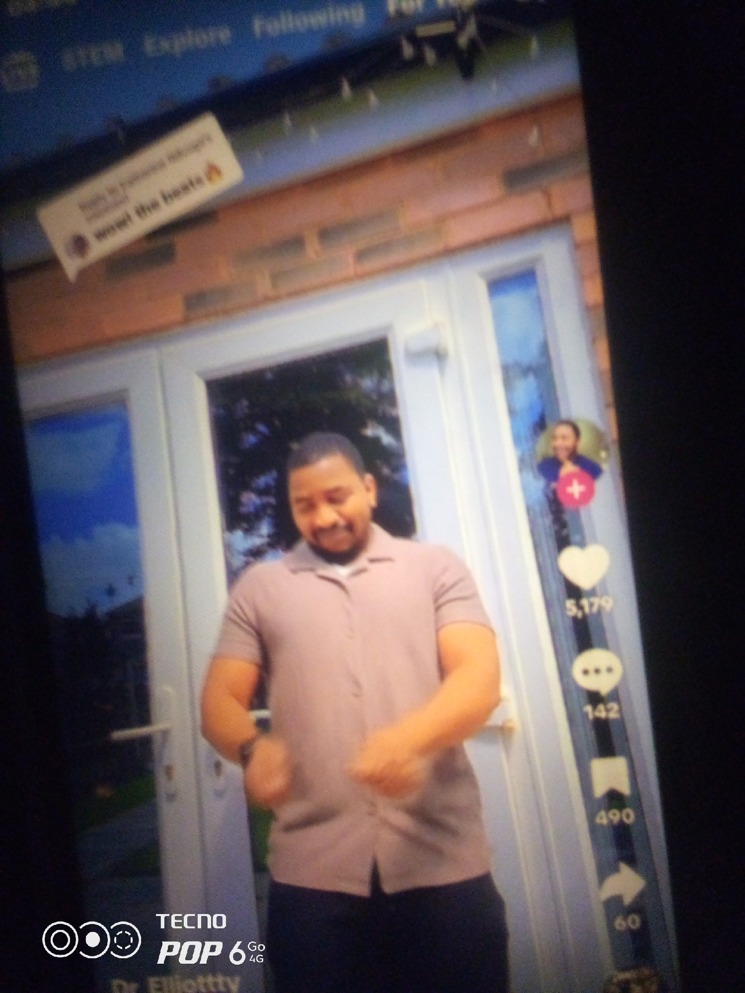 The image displays a screenshot of a social media video platform, likely TikTok, as indicated by the vertical layout of icons on the right side of the screen representing likes, comments, shares, and possibly other engagement metrics. The main subject is a man of color, appearing to be in his late 20s or early 30s, wearing a light purple polo shirt and dark pants. He is standing in front of a white door or entrance, with a brick wall visible in the background. His expression is a smile, and his body is turned slightly, suggesting he is mid-dance or posing.
In the upper left corner, a comment bubble is visible with text that reads "Wow! the beats ๐ฅ". This comment is overlaid on a blurred background that includes elements of the social media interface, such as "STEM", "Explore", and "Following".
On the right side of the screen, there are several icons stacked vertically. The topmost icon shows a circular profile picture of a person with a red plus sign overlaid, indicating a follow button. Below that, a heart icon is displayed with the number "5,179" next to it, representing likes. Further down, a speech bubble icon with three dots inside indicates comments, followed by the number "142". Below this, an icon resembling a bookmark or save symbol is shown with the number "490". Finally, an arrow icon is present with the number "60", likely representing shares or sent messages.
In the bottom left corner of the image, a logo with the text "TECNO POP 6 Go 4G" is present, along with a stylized "Dr. Elliottzy" logo.
The lighting in the image is somewhat uneven, suggesting it was taken with a phone camera, potentially in a home environment or an outdoor setting with varied light sources. The overall quality is slightly blurry, typical of a screenshot captured from a screen.
There is no specific information within the image to indicate the city of Howayi, Nigeria.