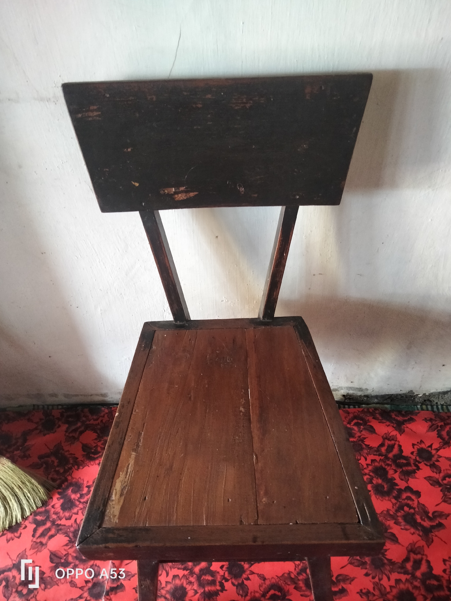 A wooden chair is positioned against a white wall in what appears to be an indoor setting in Jati, Indonesia. The chair has a dark, rectangular backrest and a brown, wooden seat. The wall behind the chair has some texture and a faint crack. The floor is covered with a red cloth that has a dark floral pattern. To the left of the chair, a portion of a broom is visible. The image also shows a watermark in the bottom left corner indicating "OPPO A53".