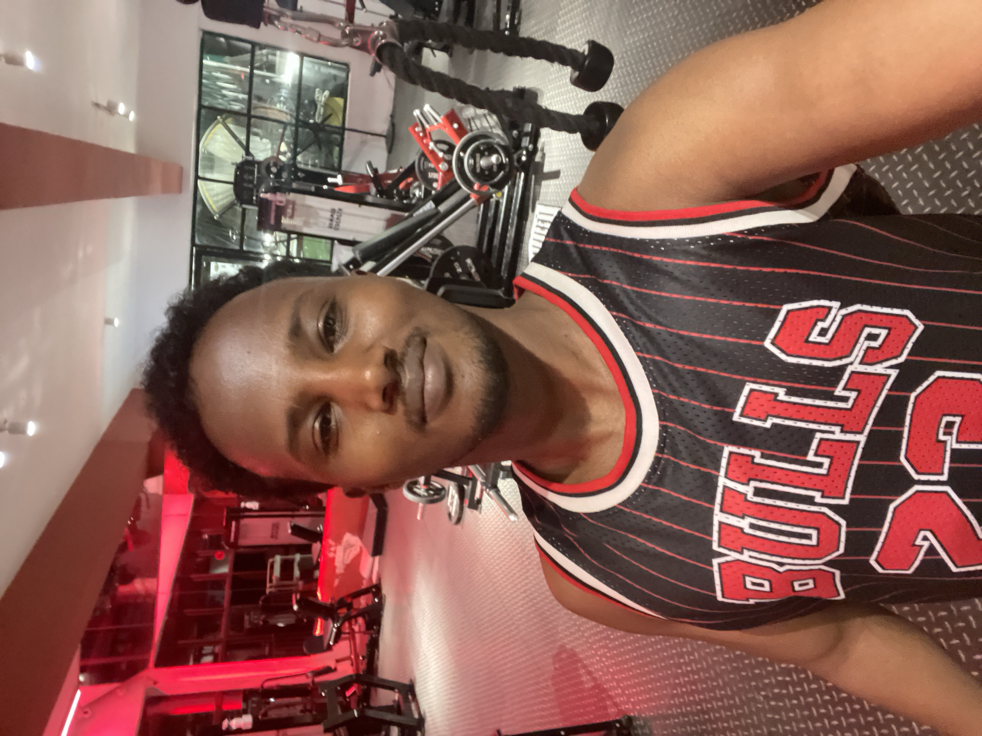 A man is taking a selfie indoors, likely in a gym. He is wearing a black and red striped Chicago Bulls jersey. Gym equipment is visible in the background. The location is consistent with a modern gym setting, but there is no definitive evidence to place it in Chongqing High-tech Zone, China.