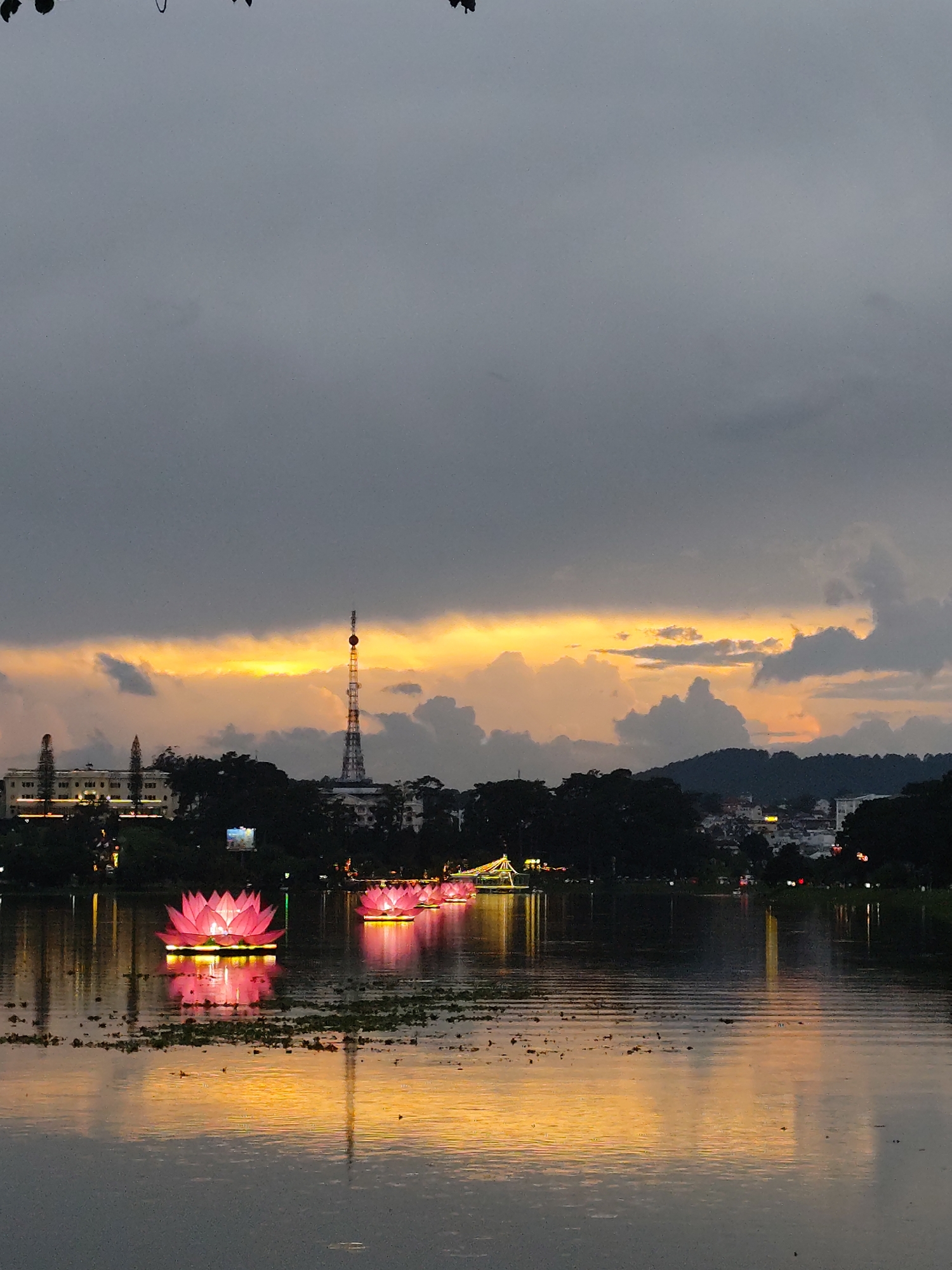 The image shows a view of Xuan Huong Lake in Da Lat, Vietnam, at dusk. The sky is cloudy with a horizontal band of orange light visible through a gap in the clouds. In the distance, the city skyline is visible with a prominent radio or television tower. Several illuminated pink lotus flower structures float on the lake’s surface. The reflections of the sky and lights are visible on the water. The foreground of the lake is darker and ripples suggest a light breeze. There are some water plants on the surface of the lake.