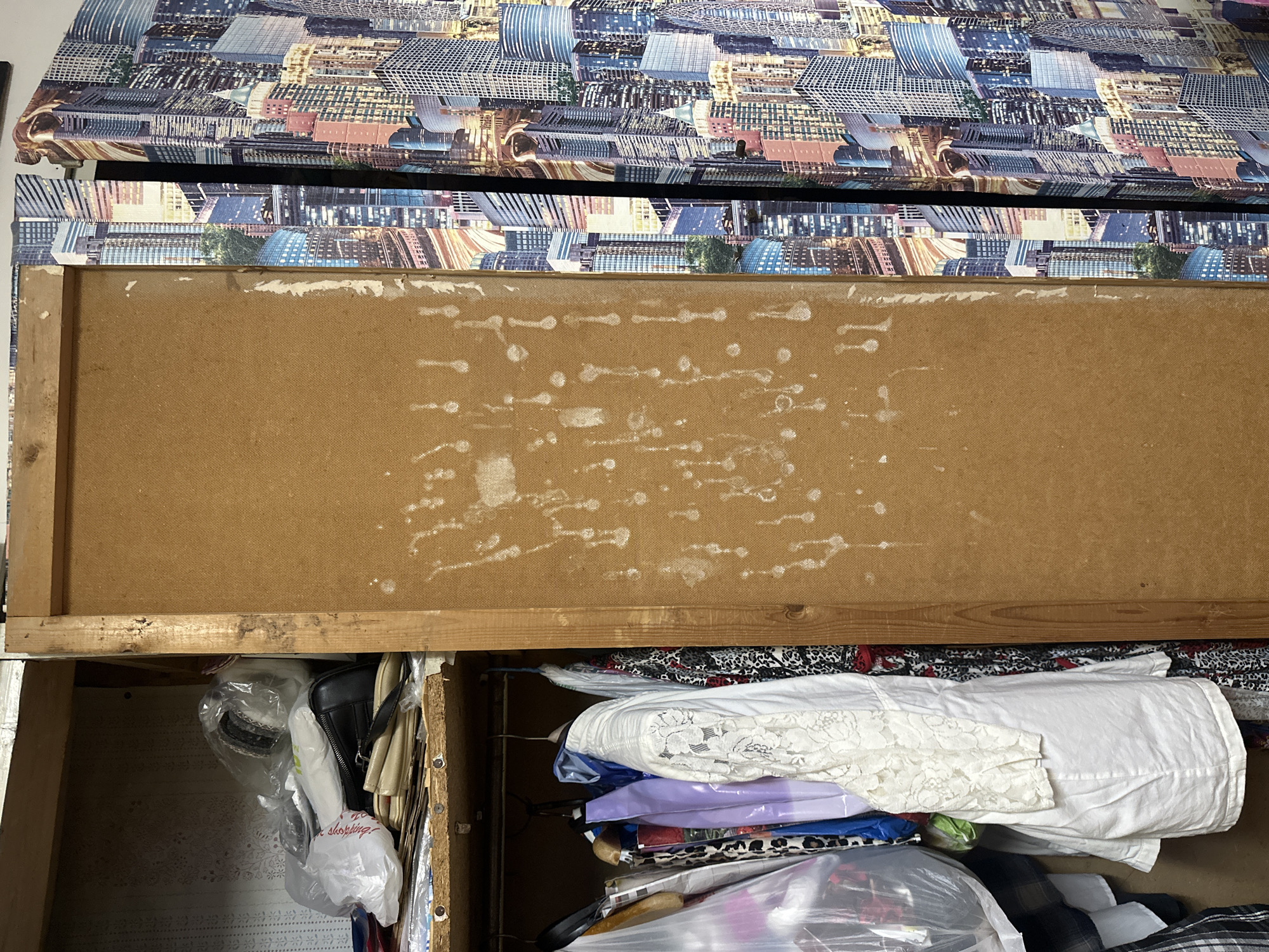 This image depicts a cluttered interior space, specifically a section of a room or closet in Aktanysh, Russia. The scene is dominated by a vertical, light brown wooden or particle board panel in the center, exhibiting numerous dried white drip marks and splatters. To the left of this central panel, an open storage unit or closet is visible. Its upper shelf holds several items, including clear and white plastic bags containing various objects, a black handbag, and other unidentified clutter, against a background of light-colored, patterned wallpaper. Below this shelf, a clothes rail supports multiple garments on hangers, including a prominent white top or dress with lace sleeves, alongside other patterned and solid-colored clothing items, some encased in plastic. To the right of the central brown panel, a vertical surface is covered with a wallpaper or fabric featuring a repetitive, collage-style print of illuminated city skyscrapers at night. A narrow dark vertical gap or seam separates this patterned surface from the central brown panel. No individuals or active interactions are present in the frame.