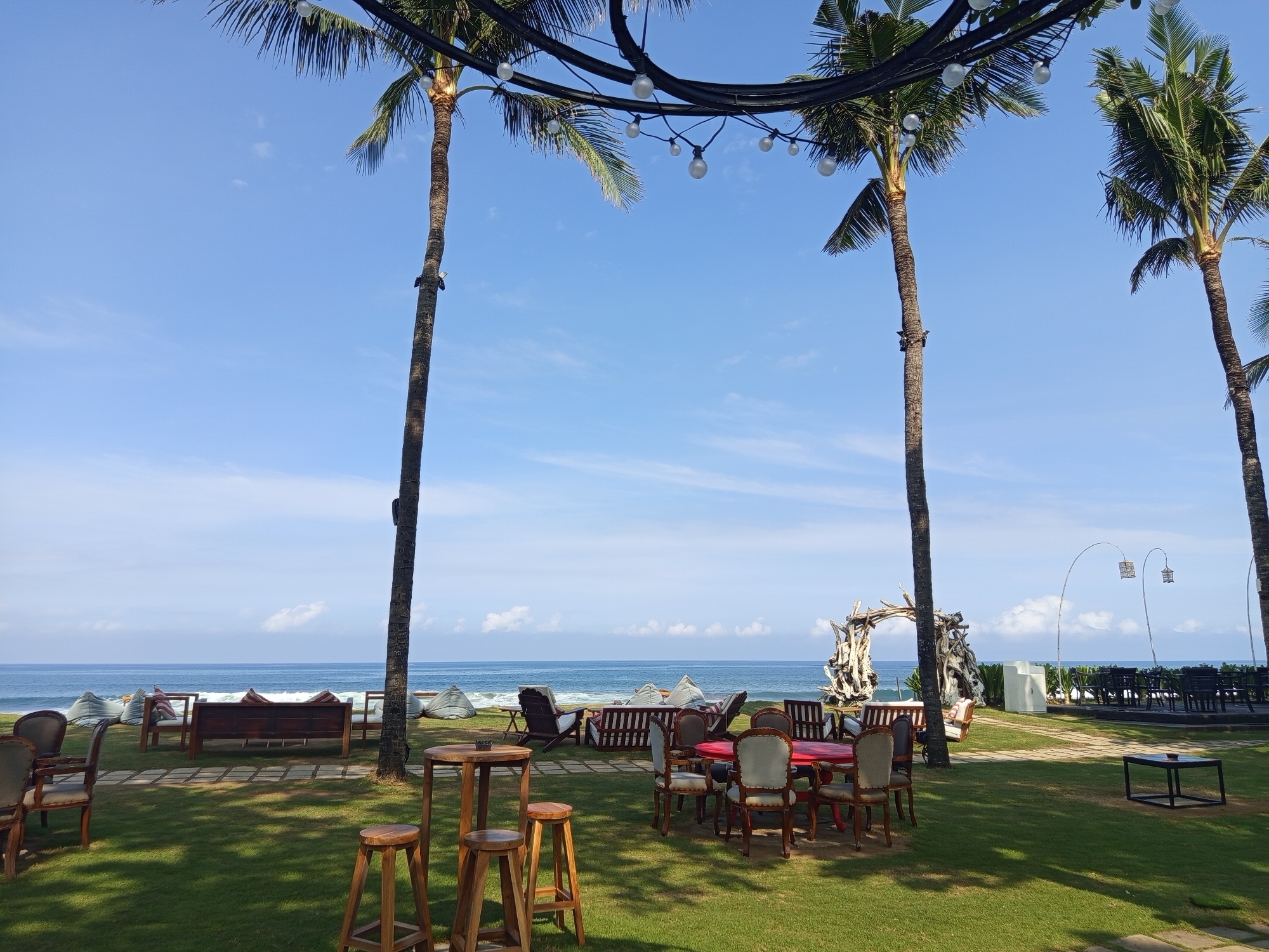 The image shows an outdoor dining area in North Kuta, Indonesia, with palm trees under which light bulbs are suspended. The foreground features a grassy lawn dotted with wooden bar stools, a red dining table surrounded by chairs, and lounge furniture. The background offers a view of the ocean with gentle waves. A driftwood structure stands near the beach. A raised platform with tables and chairs is visible to the right. The sky is clear and blue with scattered clouds.