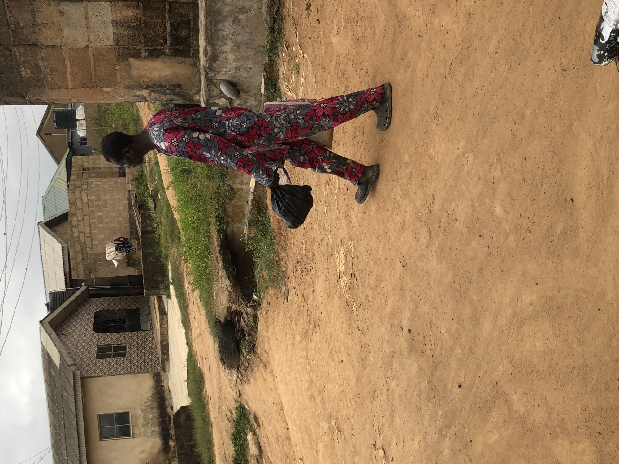 A side profile full shot shows a person walking on a dirt road in Ijoko, Nigeria. The person, wearing a brightly colored floral print outfit and dark gray Croc-style shoes, carries a black plastic bag in their right hand and a white bag is visible under their left arm. The environment consists of several buildings constructed with concrete blocks and corrugated metal roofs, some with visible water tanks. The ground is sandy and uneven, with patches of green grass and vegetation. A broken tire is present in the foreground. The sky is overcast.