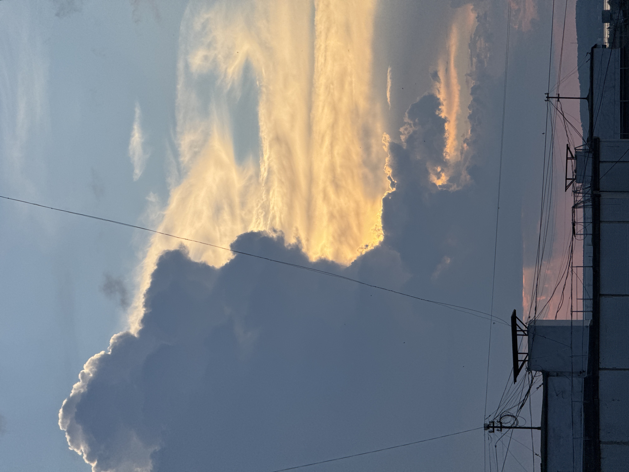 This is a medium shot, captured at eye level, of the sky above an urban area in Chaikovsky, Russia. The sky is dominated by dramatic clouds, with a large, dark cumulus cloud formation on the left, silhouetted against the bright, golden light of a setting or rising sun. The light filters through gaps in the clouds, creating streaks of orange and yellow against the blue sky. Thin power lines crisscross the frame, adding a linear element. In the foreground, the rooftops of several industrial-style buildings are visible. These structures are utilitarian, with exposed piping and electrical equipment. The overall impression is one of a busy, functional urban landscape set against a beautiful, awe-inspiring natural sky.