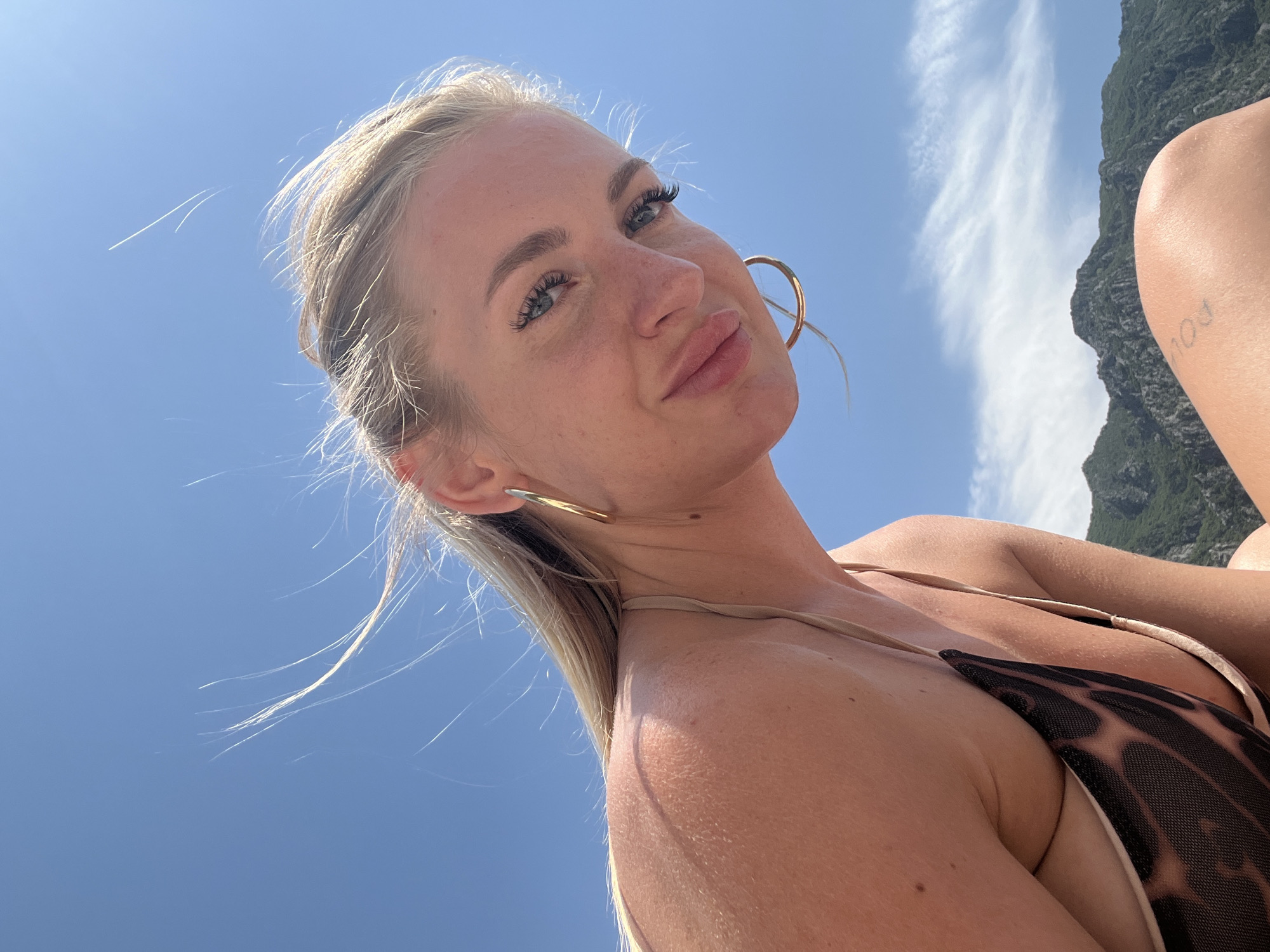 A low-angle, close-up, waist-up shot captures a blonde woman with blue eyes and light skin against a bright blue sky with wispy clouds. She is wearing a bikini with a leopard-print pattern and thin straps, and large hoop earrings. Her hair is blowing in the wind. In the background, rugged, green mountains are visible under the sky, suggesting a mountainous coastal region, possibly Risan, Montenegro, given the context. Her body is glistening with what appears to be water or sweat.