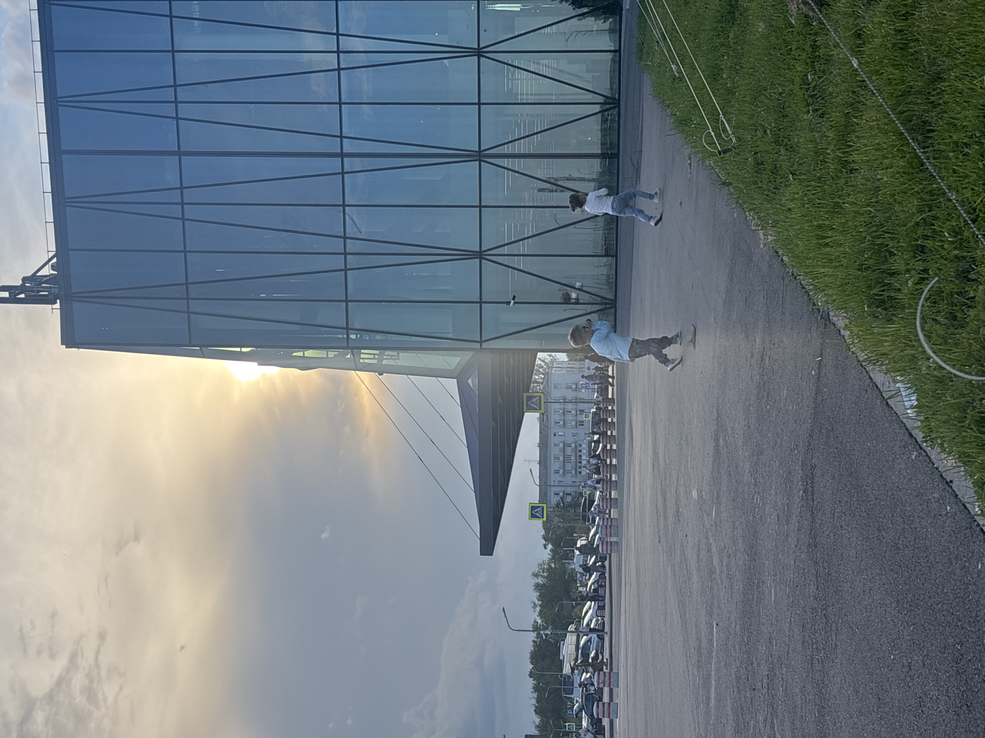 The image depicts an outdoor scene in the city of Mineralnye Vody, Russia, likely near a modern building with a glass facade. In the foreground, a paved area is visible, with green grass and wire fencing along the right edge. Two children are running on the paved surface. One child, in a blue shirt and patterned pants, is on the left, and the other, in a white shirt and blue jeans, is on the right. Behind the children, a large, contemporary building with a glass exterior and a dark, cantilevered roof structure is prominent. The sun is setting or rising, casting a bright glow through clouds on the left side of the image and reflecting on the building's glass. In the background, a parking lot with numerous cars and people, along with streetlights and some lower-rise buildings, are visible. Yellow pedestrian crossing signs are also present.