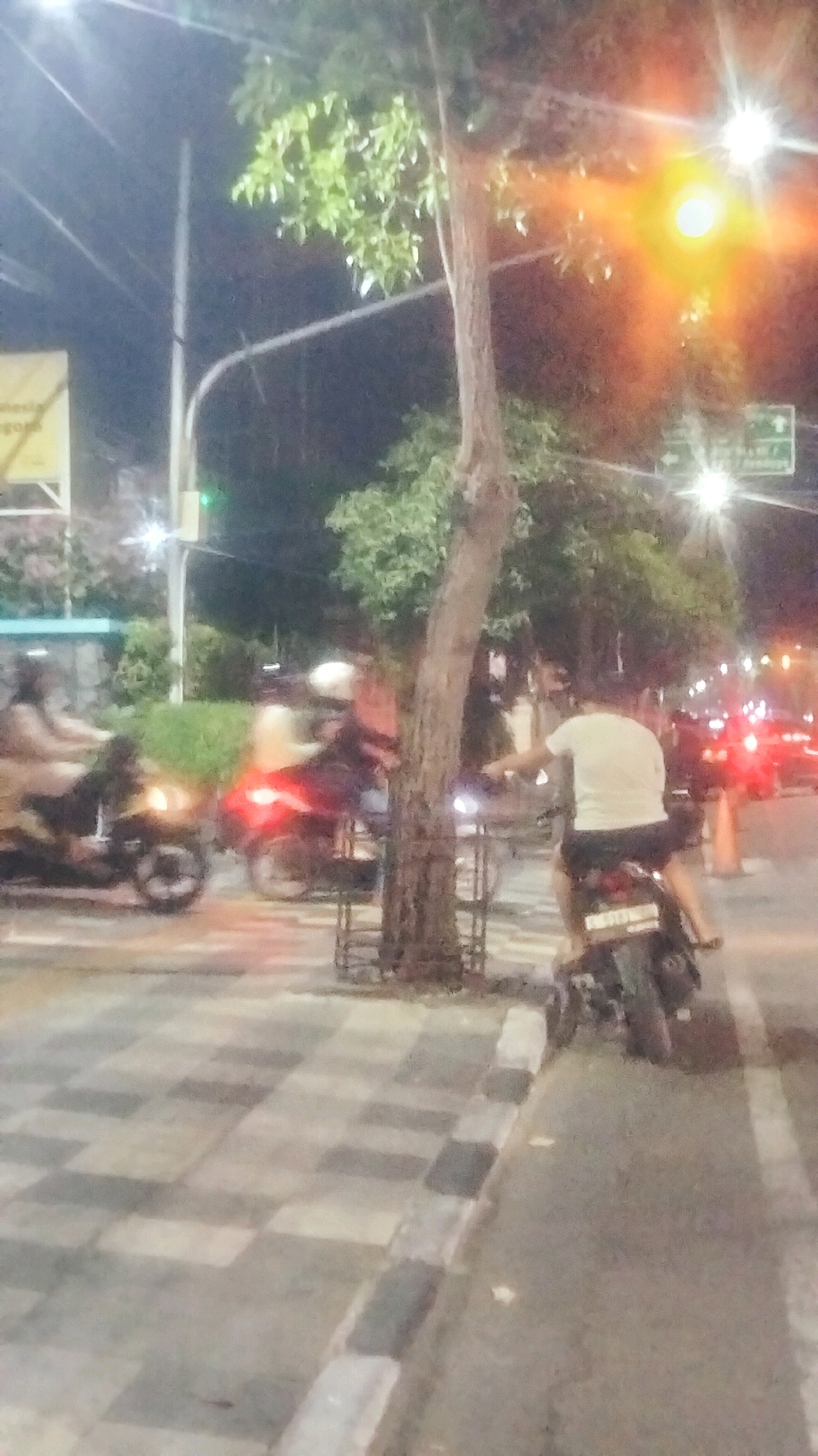 A nighttime street scene in Bojonegoro, Indonesia features a paved sidewalk with a checkered pattern of light and dark gray tiles, bordered by a curb with alternating light gray and black segments. A thin-trunked tree stands on the sidewalk, with its base enclosed by a metal grate. On the right side of the image, a person is riding a motorcycle on the asphalt road. They are wearing a white t-shirt and dark shorts, and the motorcycle has a visible license plate with the text "AG 6717 XJ". To the left of the tree, several motorcycles are also visible on the road, with one in the foreground having its red taillight illuminated, indicating it is either stopped or braking. Another motorcycle further back appears to have a rider wearing a helmet. The background is illuminated by streetlights and lights from buildings or vehicles, creating a slightly blurred and atmospheric effect, with a traffic sign visible to the right, indicating directional information.