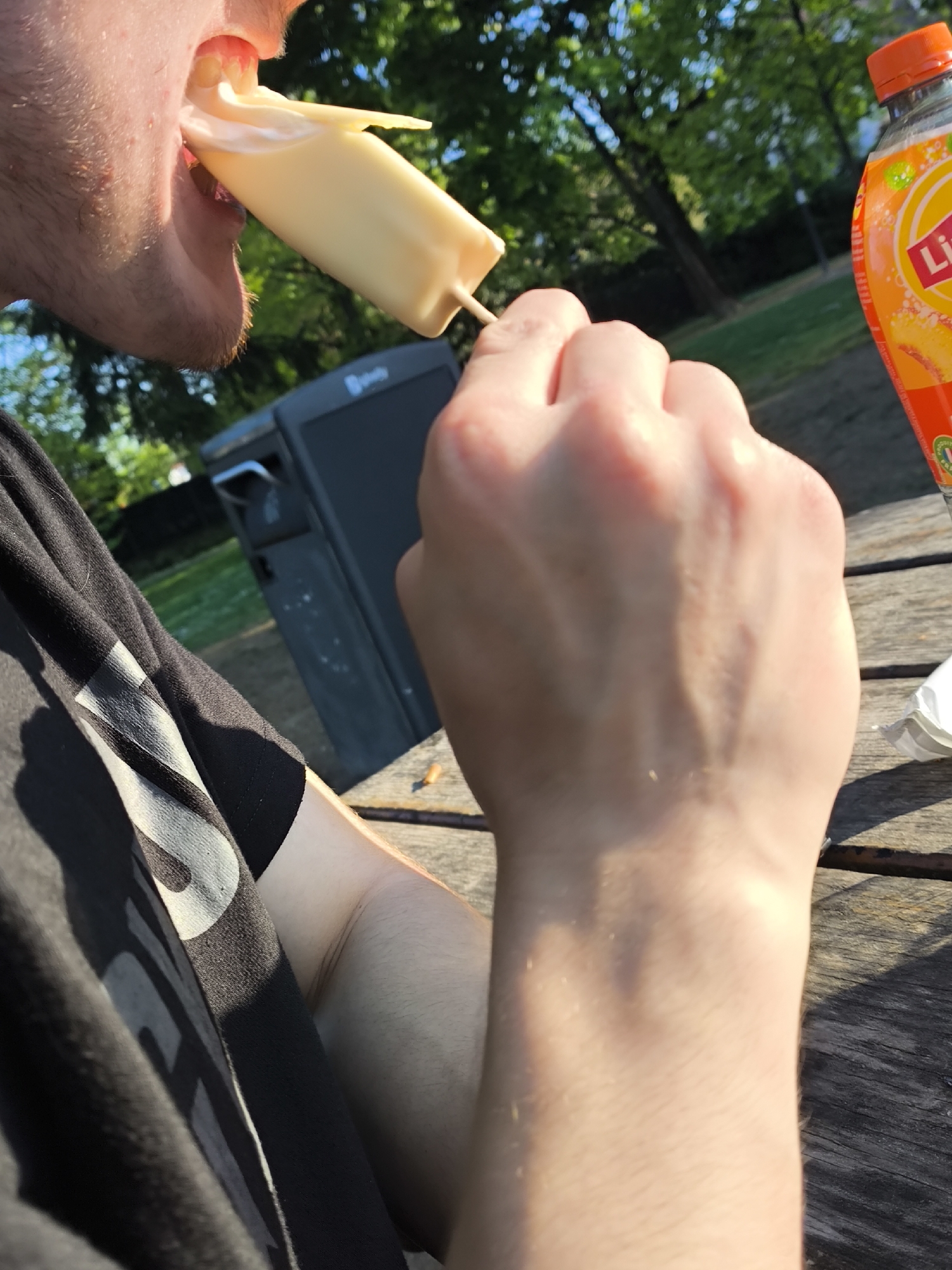 A person is eating a yellow popsicle. The person is wearing a black t-shirt. In the background, there is a black trash can, trees, and a bottle of orange soda. The scene appears to be outdoors in a park or wooded area, likely in Strasbourg, France, based on the soda brand. The person is sitting at a wooden table.