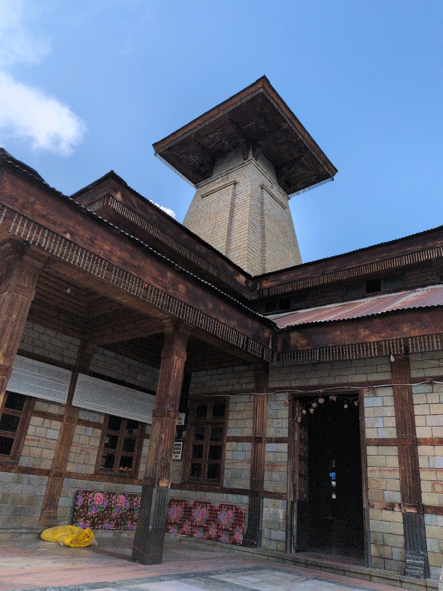 The image depicts the Hadimba Devi Temple, a Hindu temple located in the Manali city of Himachal Pradesh, India. The temple is a unique example of Himachali architecture, featuring a pagoda-style roof and intricate wooden carvings. The temple is constructed from wood and stone, with a multi-tiered roof and a tower. The surrounding area is a dense forest, and the temple is situated on a raised platform. The image captures the temple's exterior, showcasing its architectural details and its integration with the natural environment. The sky above is blue with scattered clouds, indicating a clear day. The foreground shows a courtyard area with some mats and a yellow rolled-up object.