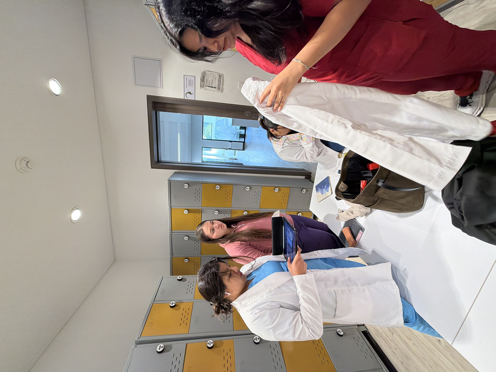 In Bojacá, Colombia, within a brightly lit, modern room featuring grey and yellow lockers lining the walls, a group of individuals, possibly medical students or professionals, are engaged in an activity around a white table. A woman in a white lab coat and blue scrubs holds a tablet, her attention focused on its screen. Beside her, a woman in a pink hoodie stands, looking in the same direction as the woman with the tablet. To the right, another woman in red scrubs is holding up a white garment, seemingly examining it. Further in the background, through an open doorway, a hallway is visible with additional rooms. The environment suggests a locker room or changing area, possibly within an educational or healthcare facility.