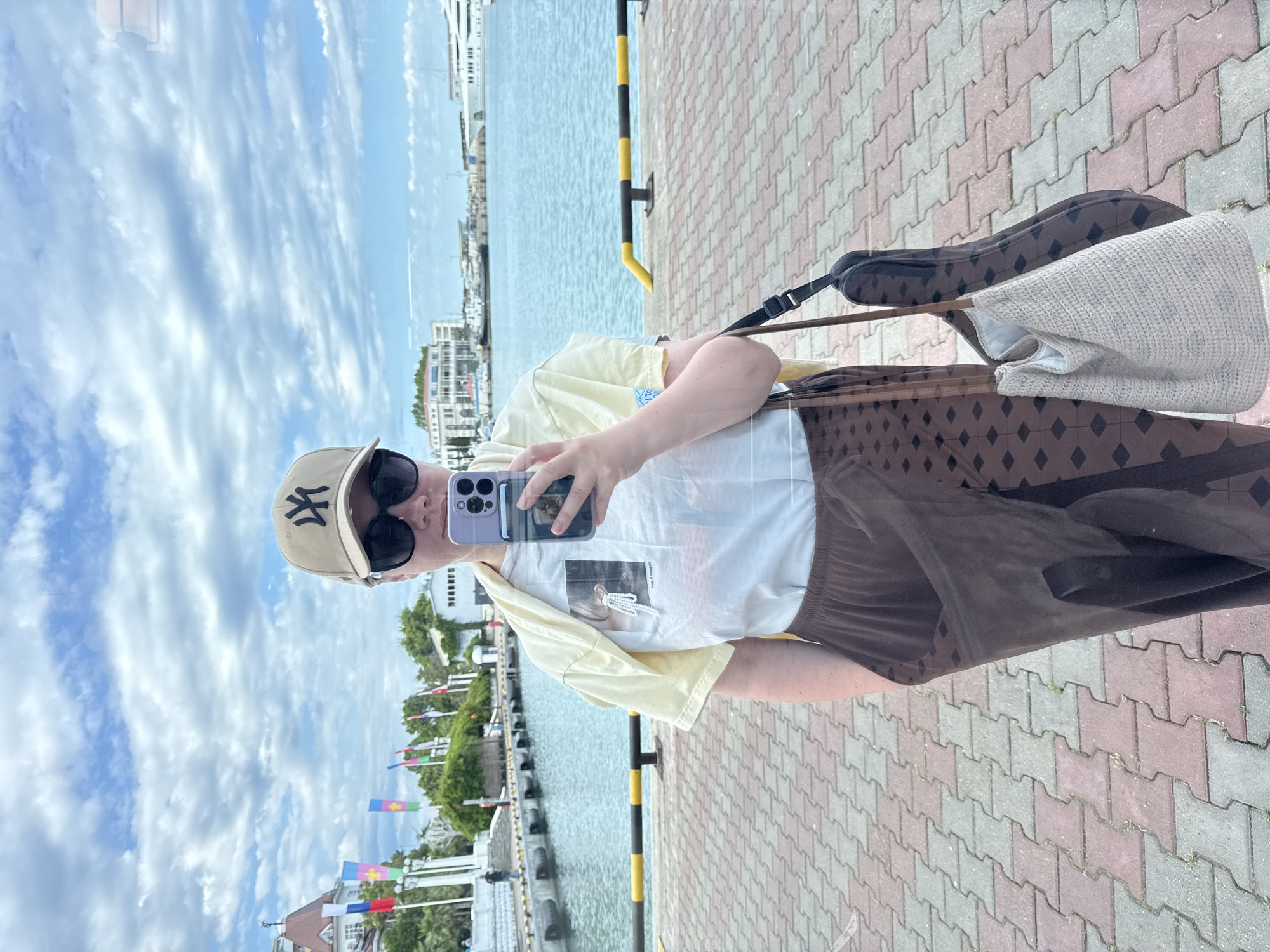 A woman stands on a paved waterfront in Sochi, Russia, taking a selfie with her phone. She is wearing a beige baseball cap with a black logo, sunglasses, a light yellow t-shirt layered over a white graphic t-shirt, and brown patterned pants. A light-colored woven bag hangs from her left shoulder, and a dark guitar case is slung behind her. The background shows the turquoise waters of the sea, a promenade lined with flags and trees, and buildings in the distance, characteristic of Sochi's coastal architecture. The sky is cloudy with patches of blue.