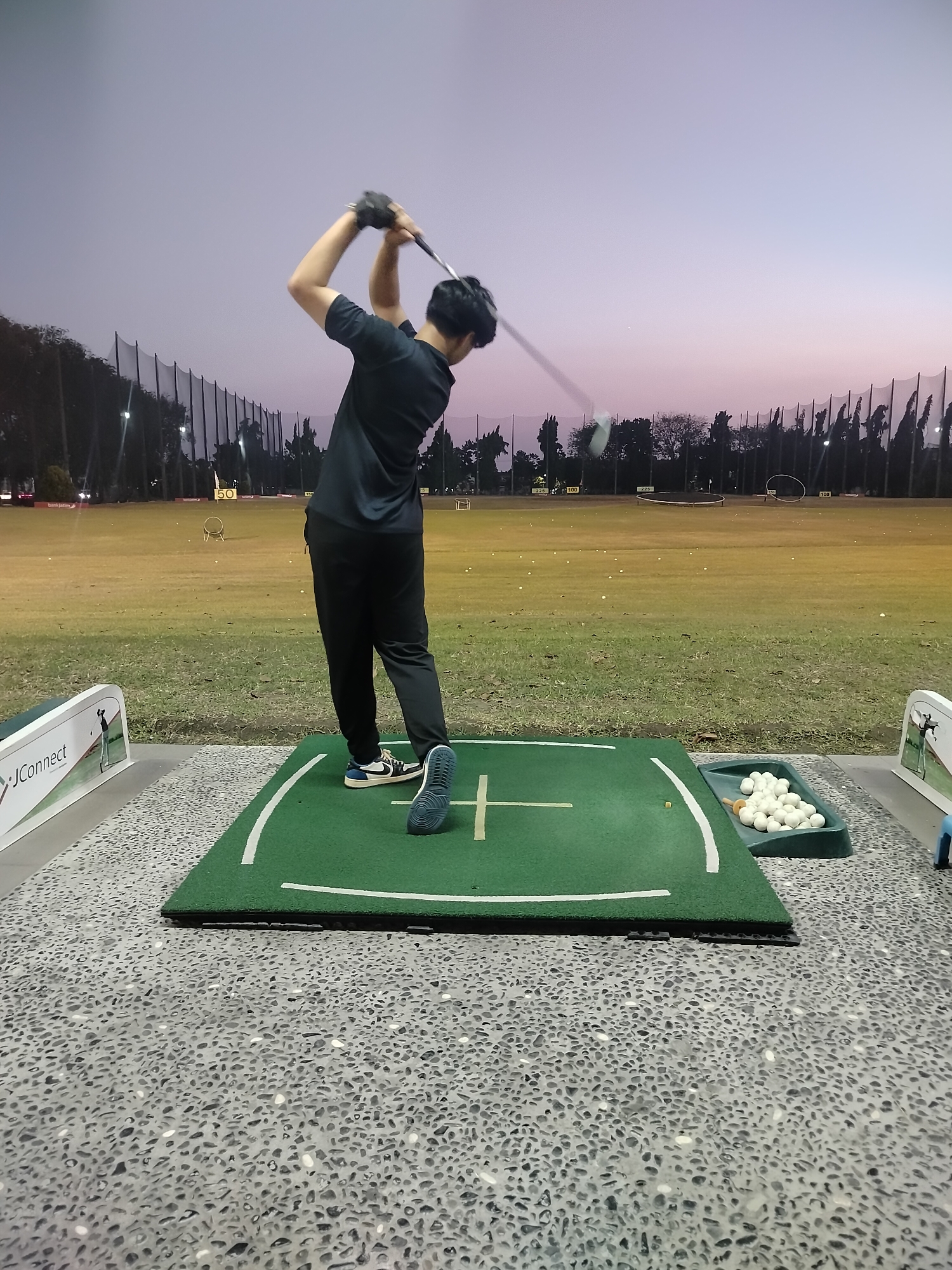 A person is playing golf at a driving range in Surabaya, Indonesia, during twilight. The person is wearing a black t-shirt and black pants, and is in the process of swinging a golf club. They are standing on a green golf mat with white markings, and there is a tray of golf balls next to the mat. In the background, there is a large, grassy field with many golf balls scattered on it. There are also golf nets and trees in the distance. The sky is a gradient of purple and pink, indicating sunset or sunrise.