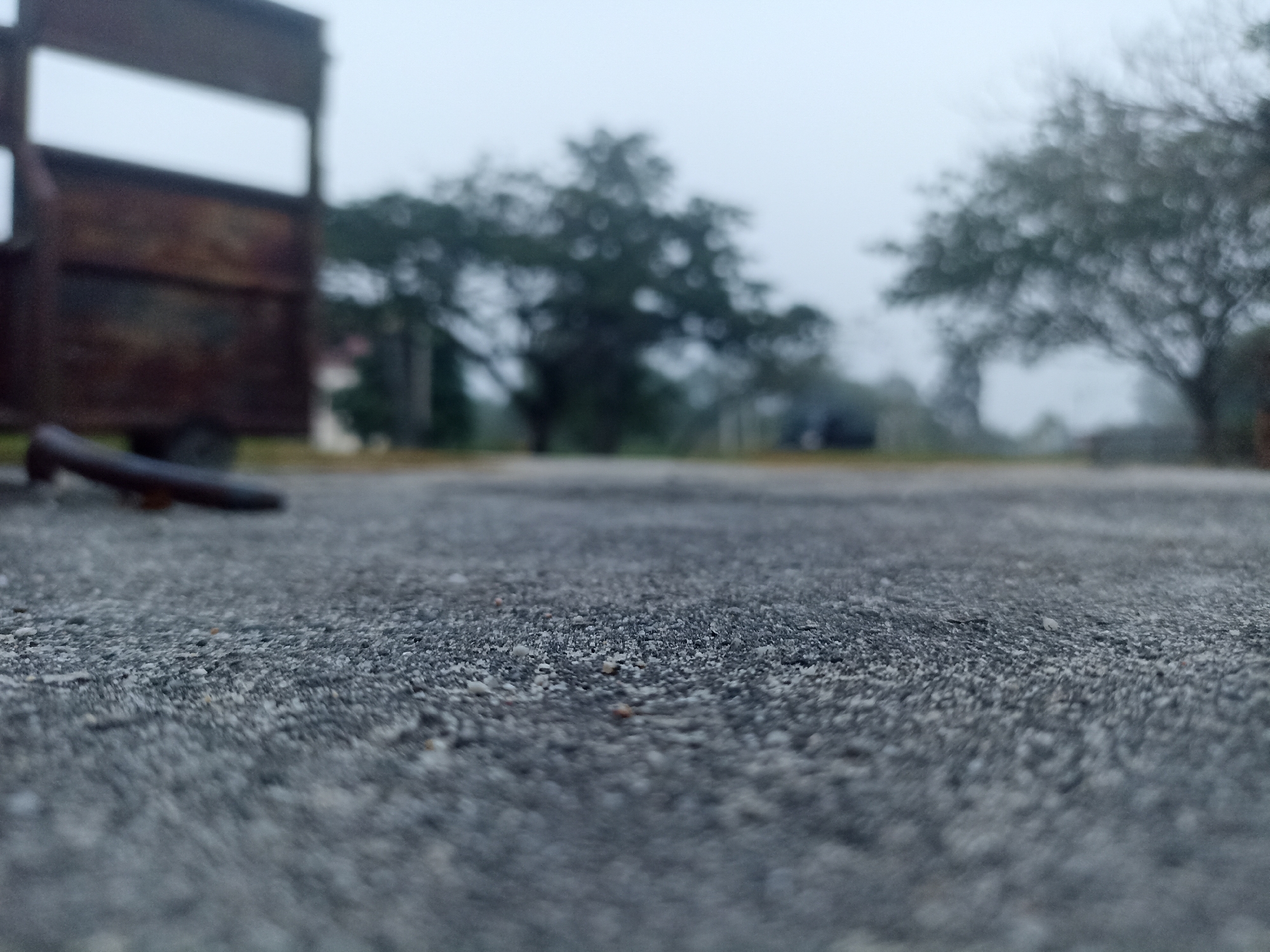 The image depicts a low-angle view of a weathered asphalt surface. In the background, out of focus, is a dark, rustic-looking metal structure, possibly part of a trailer or cart, positioned to the left. Further in the distance, blurred trees with dark foliage are visible against a hazy, overcast sky. The scene appears to be an outdoor location, possibly a rural or undeveloped area. No people or specific actions are discernible in the image. The environment suggests a calm, possibly damp atmosphere due to the muted colors and lack of strong sunlight. The specific location of Kampung Tengah, Indonesia, is not directly evident from the visual information presented in the image itself.