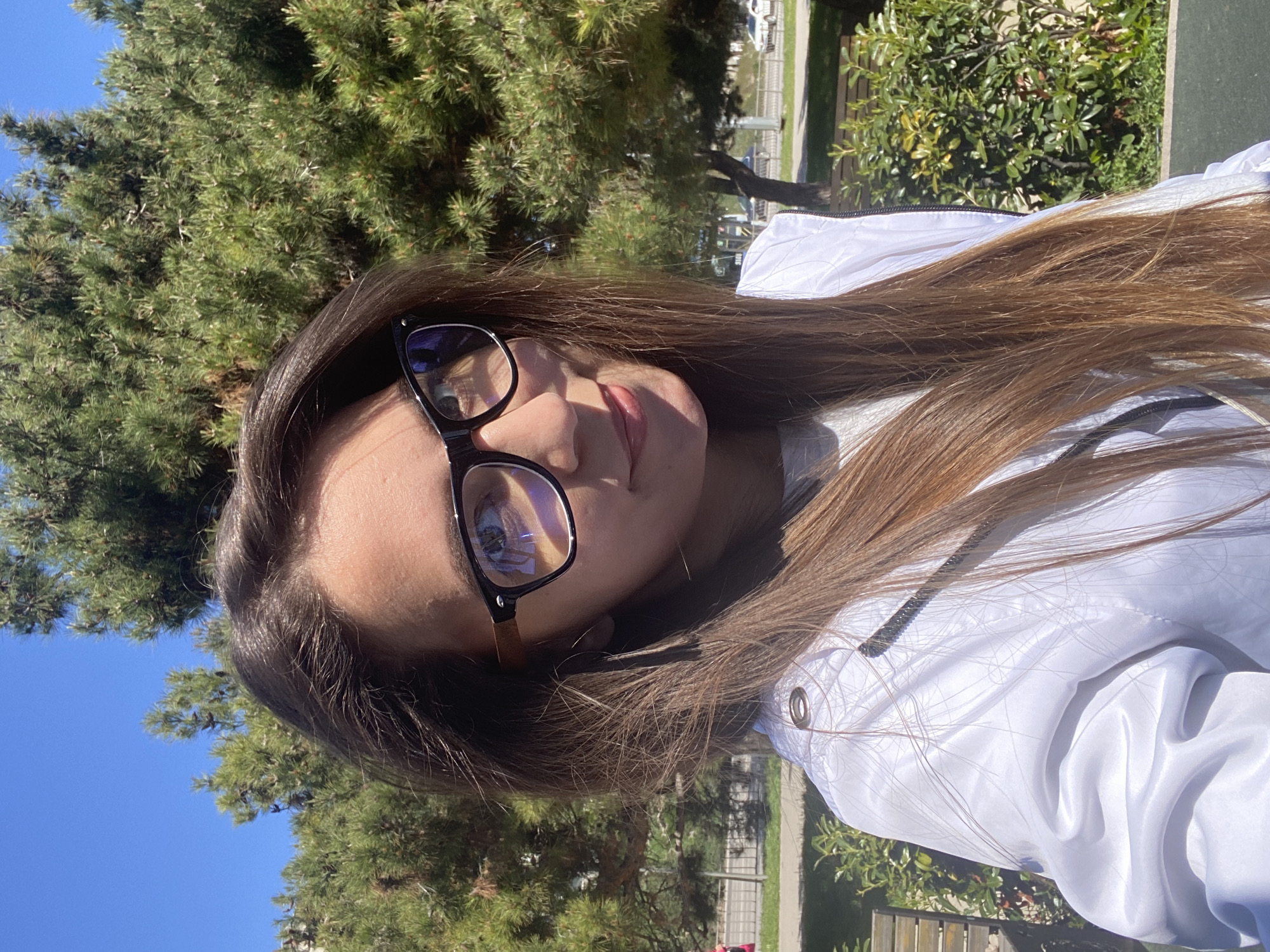 A young woman wearing glasses and a white jacket stands outdoors in what appears to be a park-like setting. The background includes trees and a clear blue sky. The photo appears to be taken at an angle. There is no specific information to confirm that this image was taken in Sevastopol, Ukraine.