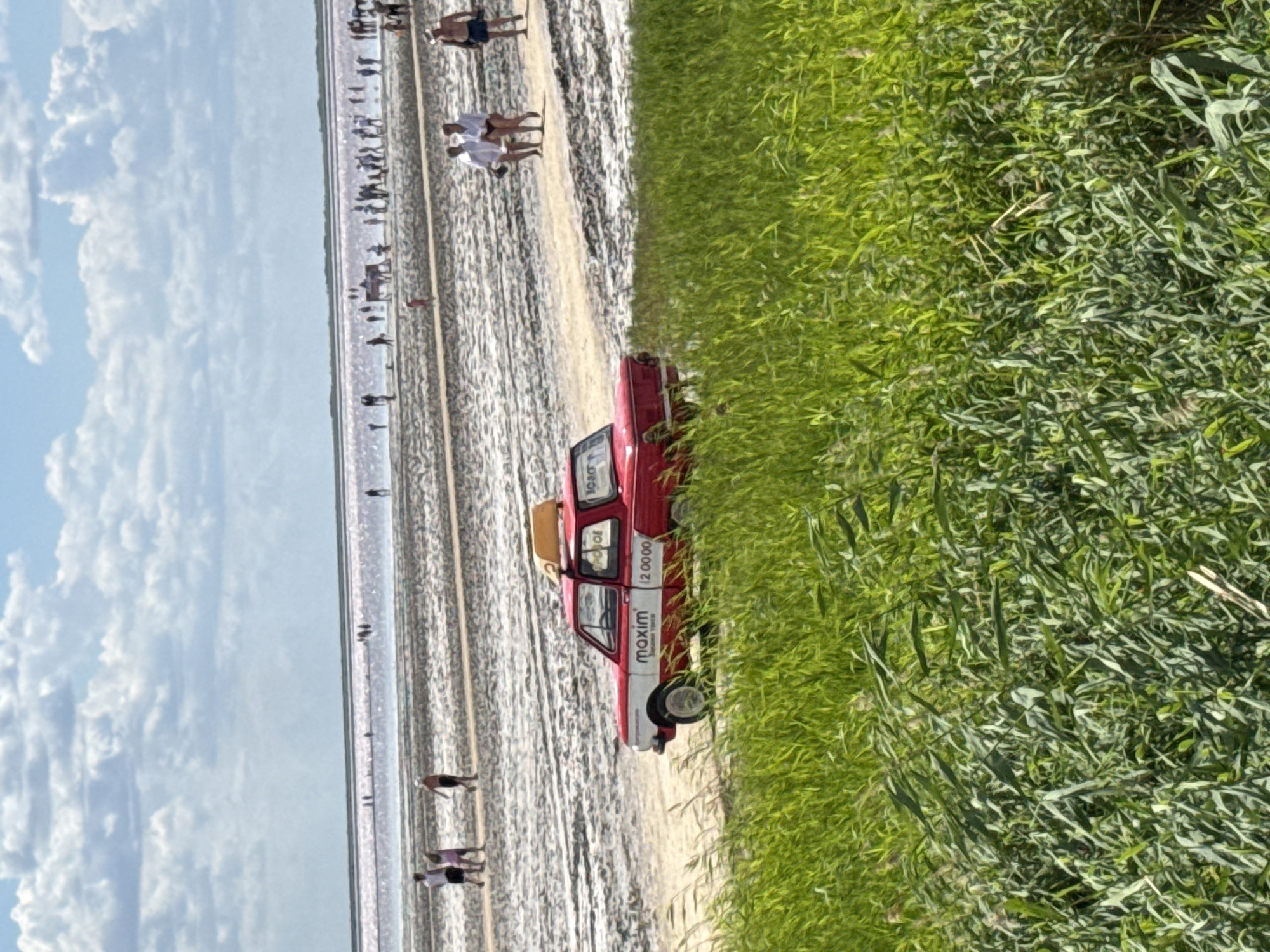 In Бурсоль, Russia, a vintage red and white taxi with a yellow sign on its roof drives along a dirt path on a salt lake. The car displays the Maxim logo and the digits "12 00 00". Several people are walking in the distance on the salt flats towards the pink-hued water. Tall green grass dominates the foreground. The sky is partly cloudy.