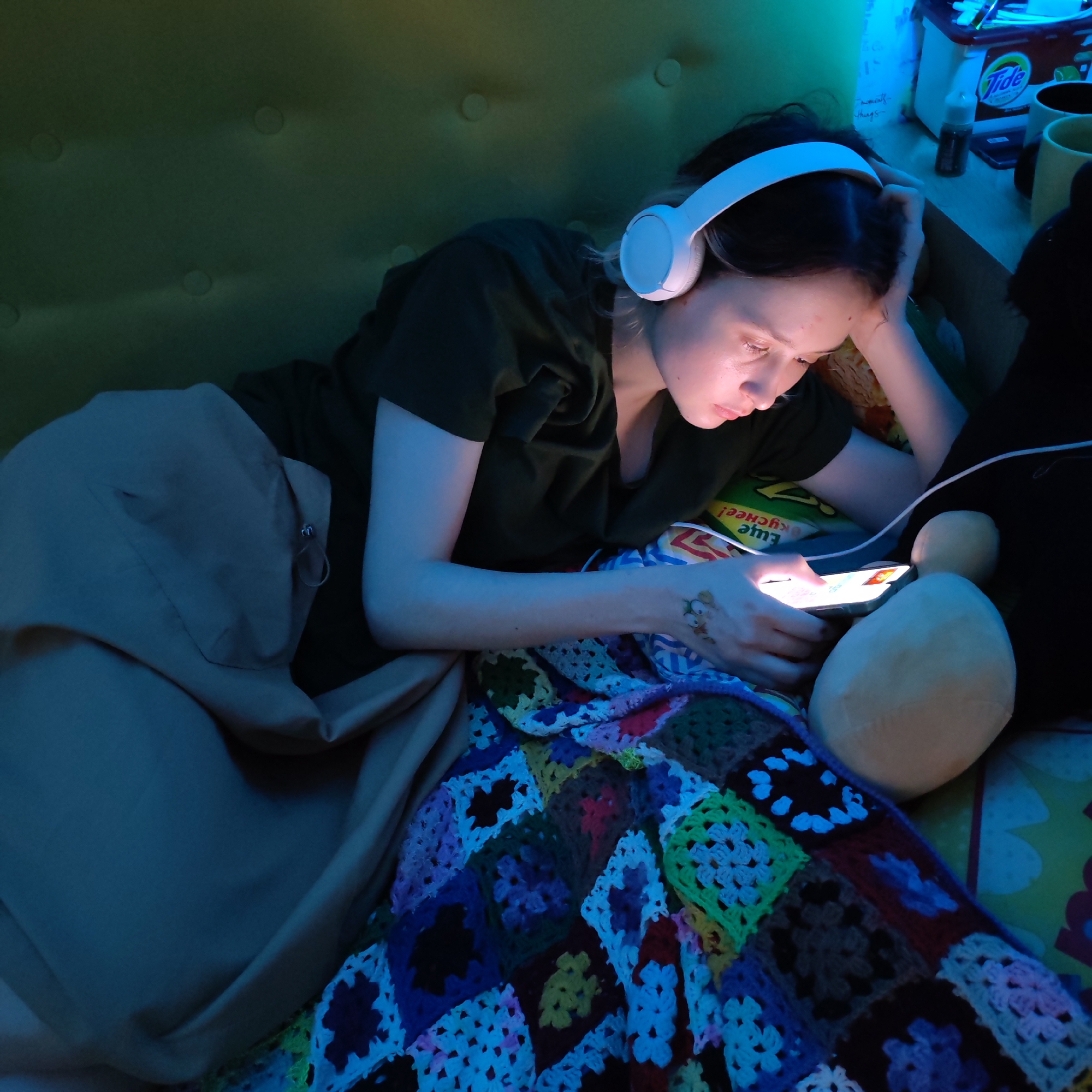 A young woman is lying on a green tufted sofa or bed, covered by a light brown blanket and a colorful crocheted quilt. She is wearing white headphones and a dark green T-shirt, and she is looking at a smartphone in her hands. Her right arm rests on the sofa, with her hand near her head. A large stuffed animal is next to her on the right side. A table or shelf is visible behind her head, with various items on it including a container of Tide detergent, mugs, and other unidentified objects. The scene is lit with a blue light, casting an overall blue hue on the image. The location cannot be determined based on visual information.