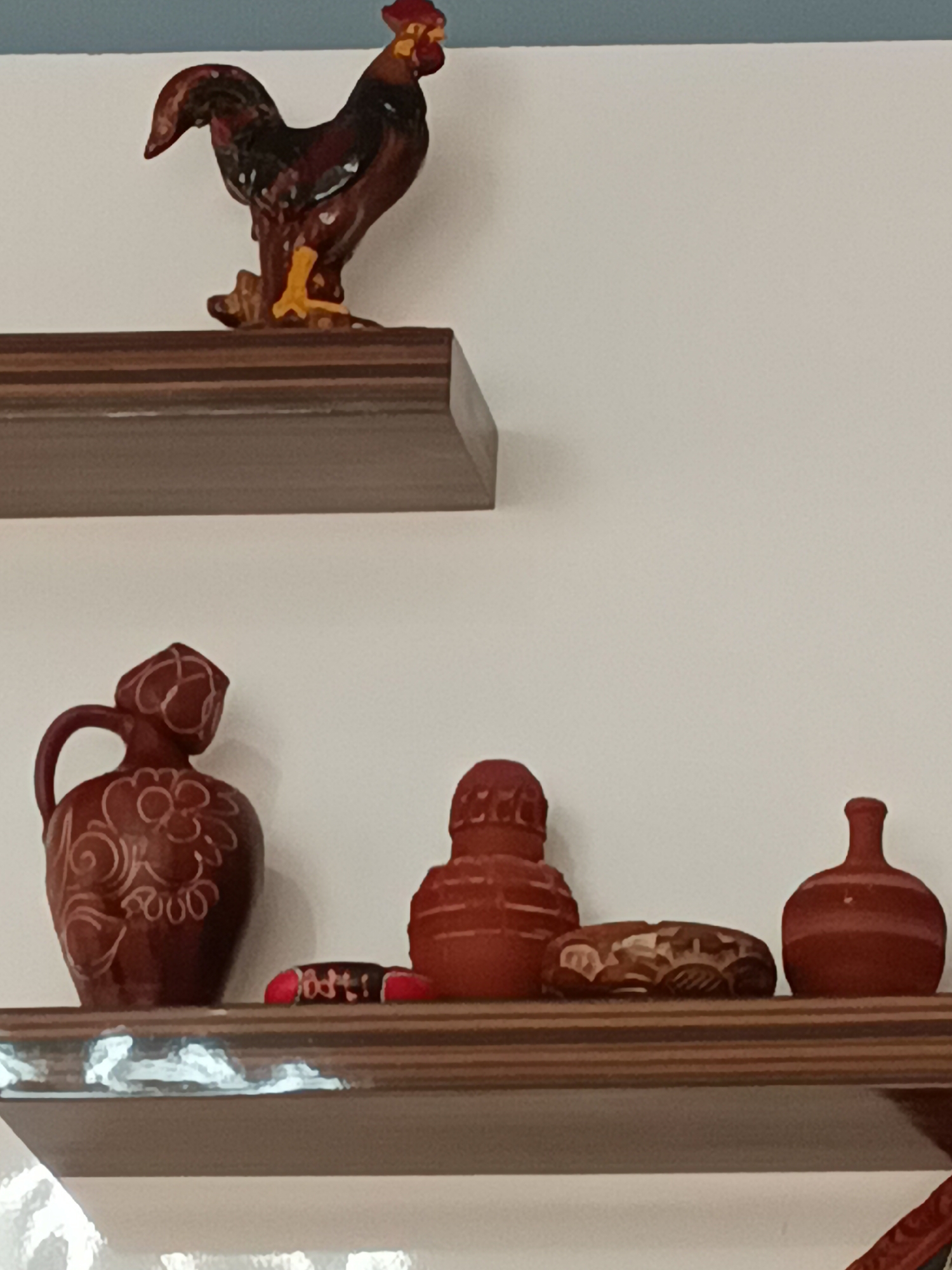 This media file displays a close-up, indoor shot of two wooden shelves mounted on a white wall. The shelves are positioned one above the other. The upper shelf holds a decorative ceramic rooster, predominantly brown with red and yellow accents, standing on a brown base. The rooster is depicted in a proud stance, with its tail feathers raised.
The lower shelf showcases a collection of small, reddish-brown ceramic items. From left to right, there is a small jug with a handle and a decorative floral pattern in a lighter color, followed by a cylindrical object with ribbed detailing. Next to it is a carved, oval-shaped object with intricate patterns. Finally, on the far right, is a small, bulbous vase with a narrow neck. Between the jug and the cylindrical object, a small, cylindrical item with red and black markings is visible, possibly a decorative stick or a small sculpture. The shelves themselves have a wooden veneer with some visible wear or chips, particularly on the lower shelf. The lighting suggests an indoor setting, possibly during the day, as there are no strong shadows. The overall scene is one of decorative display, with a rustic or traditional aesthetic. There is no visible text. The implied location context is Alanya, Turkey.