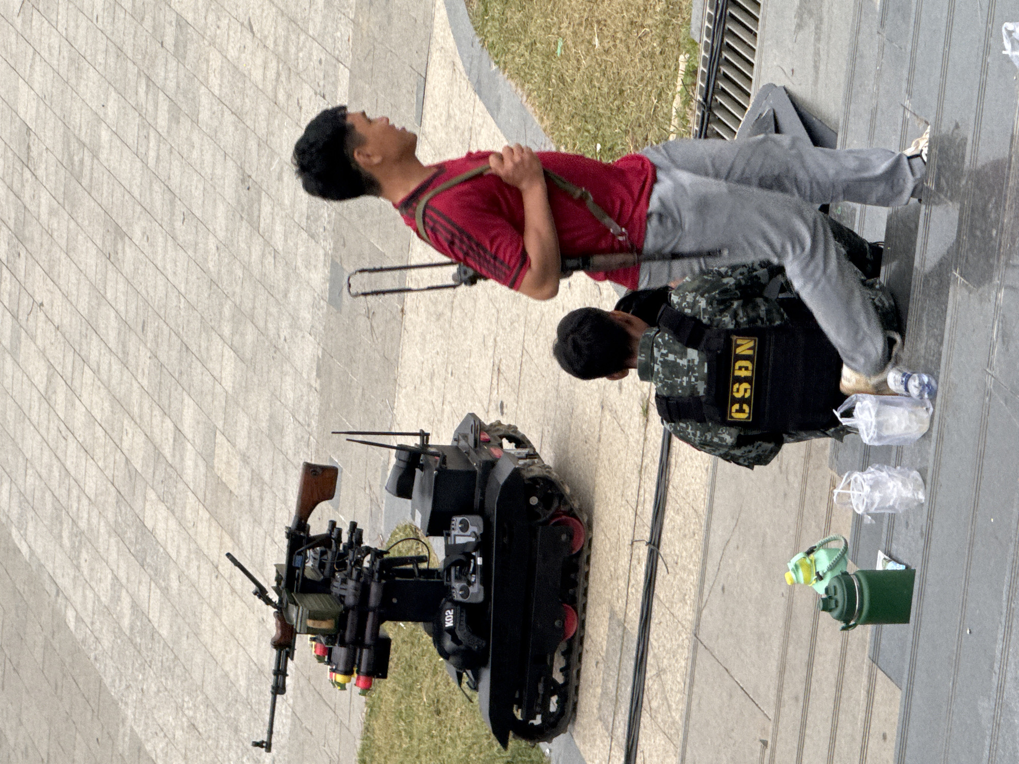 The image depicts a street scene in Da Lat, Vietnam, featuring a remote-controlled robot equipped with a machine gun and what appear to be rockets. The robot is positioned on a tiled surface, with a grassy area to its left. In the foreground, two individuals are present. One person, wearing a camouflage vest with the letters "CSDN" visible, is kneeling on a set of stairs. Another person, wearing a red t-shirt and gray pants, walks up the stairs carrying a rifle slung over their shoulder. Several plastic bottles and a green thermos are placed on the stairs. The background shows more tiled pavement. The overall scene suggests a security or public safety operation in progress.