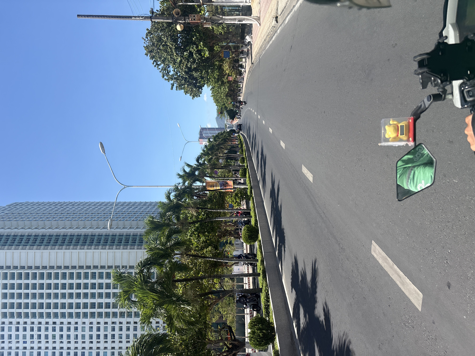 The image shows a road in Nha Trang Ward, Vietnam, flanked by palm trees and a tall, white, grid-patterned building. A motorcycle, partially visible in the foreground, has a small, yellow toy in a clear plastic box attached to the handlebars. The road stretches into the distance, marked by white lines. Other motorcycles and trees line the roadside. The sky is clear and blue. The image appears to have been taken from the perspective of the person operating the motorcycle.
