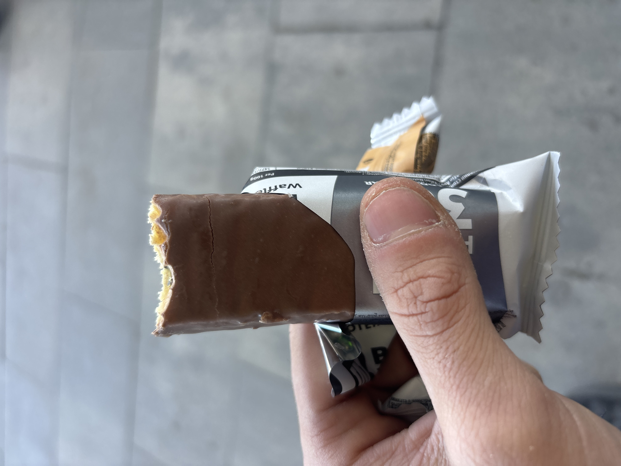 A hand holds a partially eaten chocolate-covered wafer bar in its opened packaging. The packaging is primarily white and gray with black lettering. The bar is brown with a visible wafer interior at the bite mark. The background is a blurred gray, suggesting an urban setting. Without additional context, it's not possible to confirm this scene is located in Plovdiv, Bulgaria.
