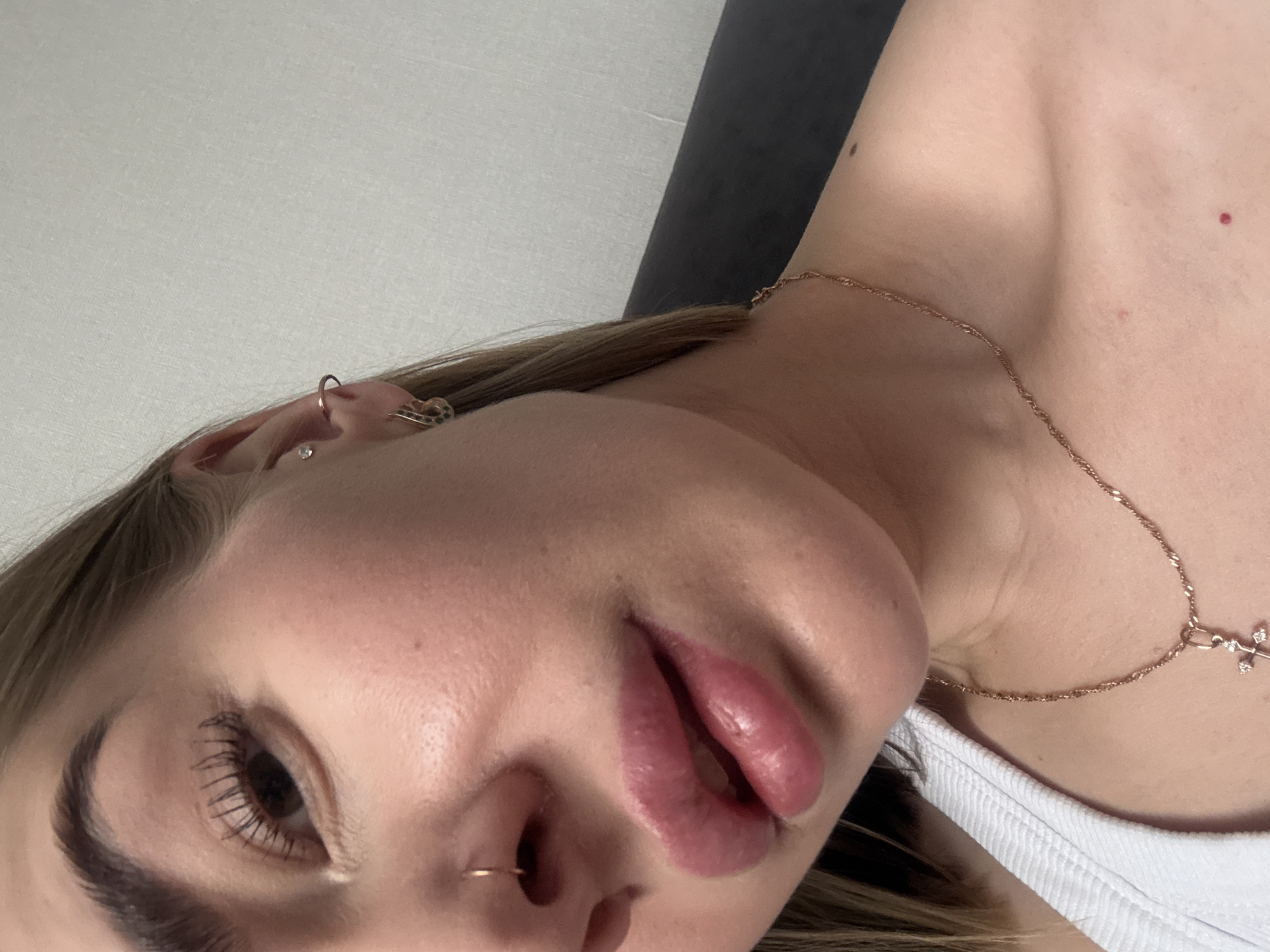 A close-up, angled shot of a woman's face and upper torso. She has light skin, brown eyes, and fair hair. Her lips are a reddish-pink color. She has a nose ring and multiple earrings in her left ear. She is wearing a white top and a delicate gold necklace with a cross pendant. There are small moles visible on her chest and neck. The background is a light gray wall and a dark gray cushion or headboard. The location in the city of Chaikovsky, Russia is not discernible from the image.