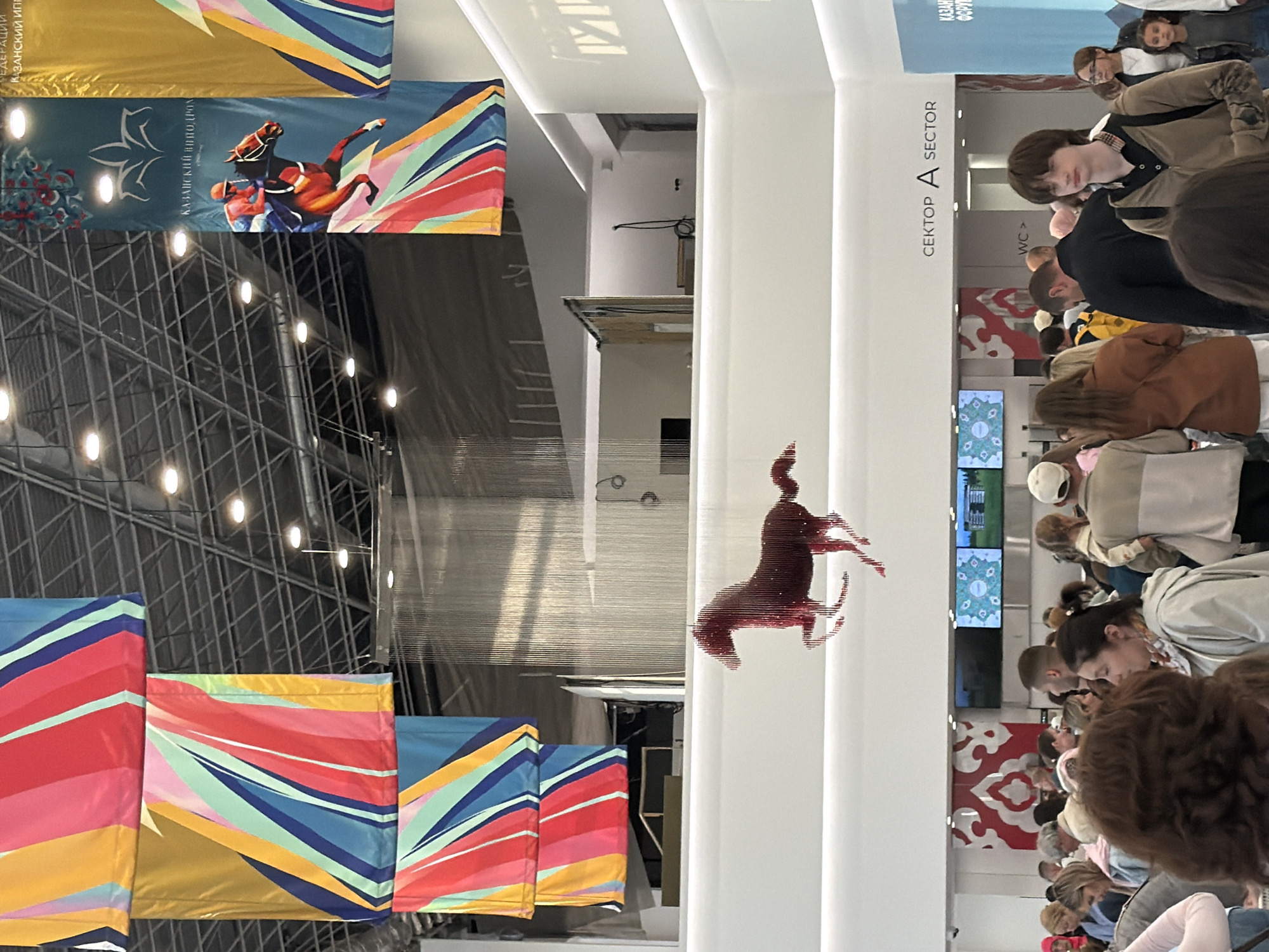 The image shows a gathering of people indoors, likely at an exhibition or event. Several large banners with colorful abstract designs are displayed. One banner features a stylized image of a horse and rider with intricate patterns. In the background, there is a metal grid structure with lights. A vertical white pillar has text labels "SEKTOR A" and "SECTOR". Further down the pillar, a sign reads "WC".
A unique art installation of a red horse silhouette made of small pieces is suspended from the ceiling. Several people are standing and walking around, appearing to observe the displays or move through the space. Some individuals are looking at screens mounted on the wall, which show various graphics and images. The lighting suggests an indoor environment, possibly well-lit with artificial lights. The mood seems neutral, with people engaged in their activities. The overall setting appears to be a modern, spacious venue. The banner text is in Russian.