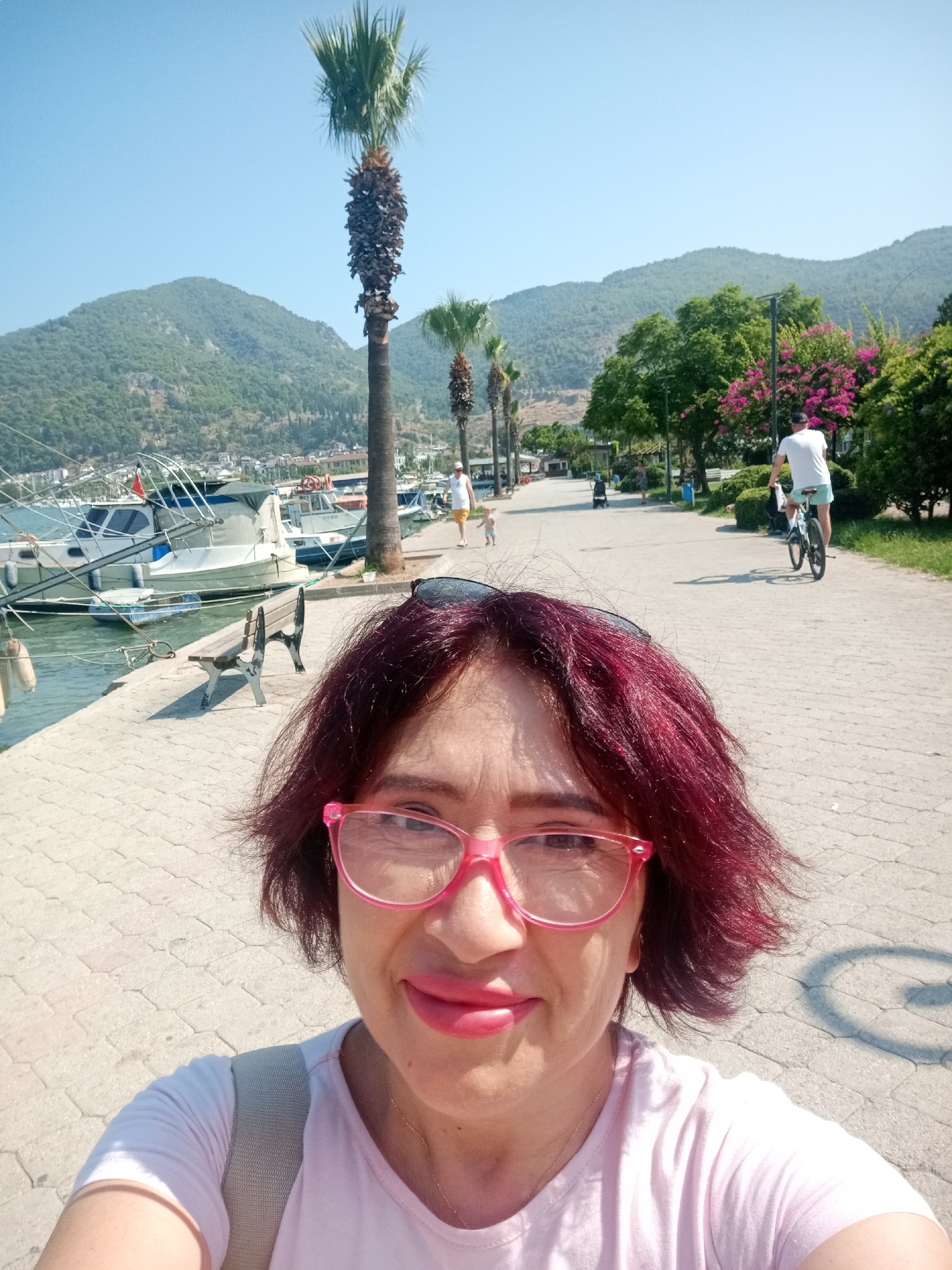 A woman with short, dark red hair and pink glasses takes a selfie on a paved walkway by the water. She is wearing a light pink t-shirt and a grey backpack strap is visible. Behind her, boats are docked on the left, and a walkway with trees and mountains in the background is visible on the right. A man rides a bicycle on the walkway, and another person walks in the distance. The location appears to be a waterfront promenade in Fethiye, Turkey, characterized by its palm trees, boats, and mountainous landscape.